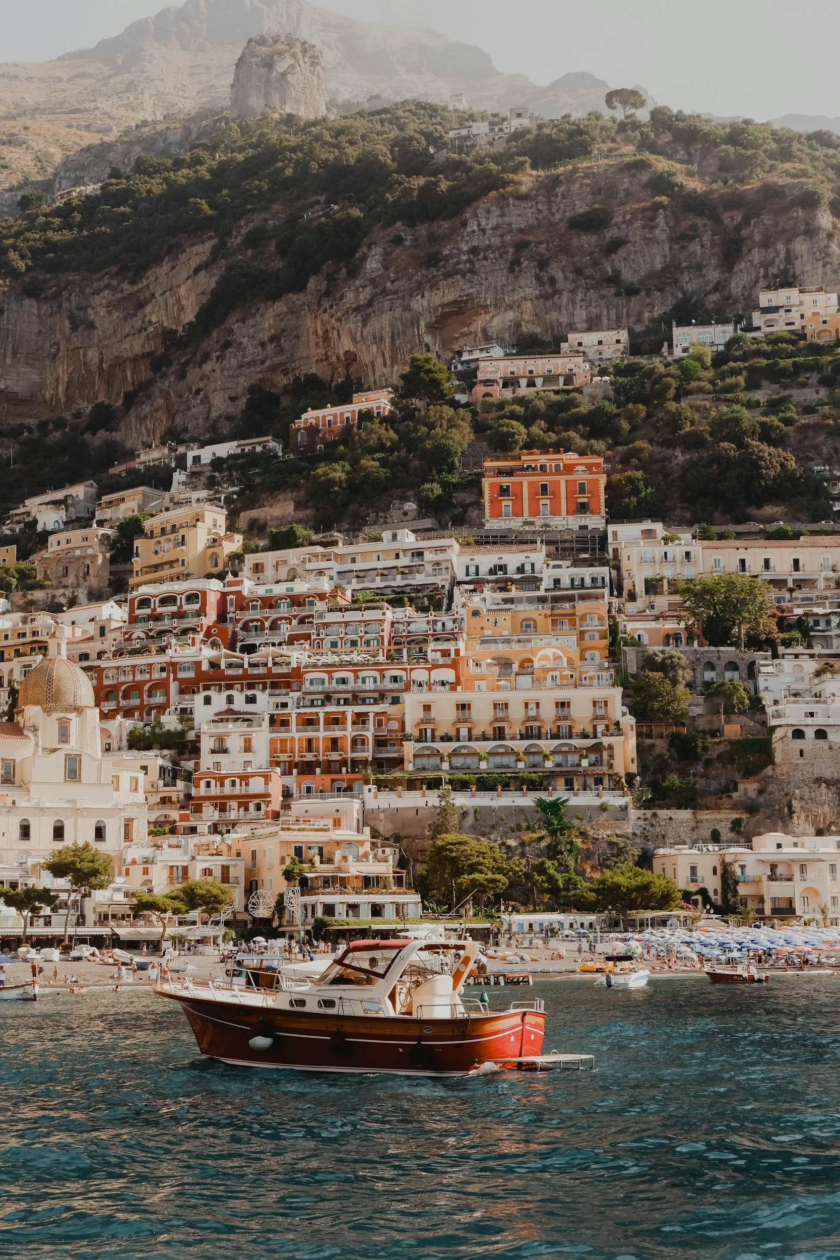 A coastal scene with a boat docked in water, in front of a hillside filled with colorful buildings and green trees, and mountainous terrain in the background.