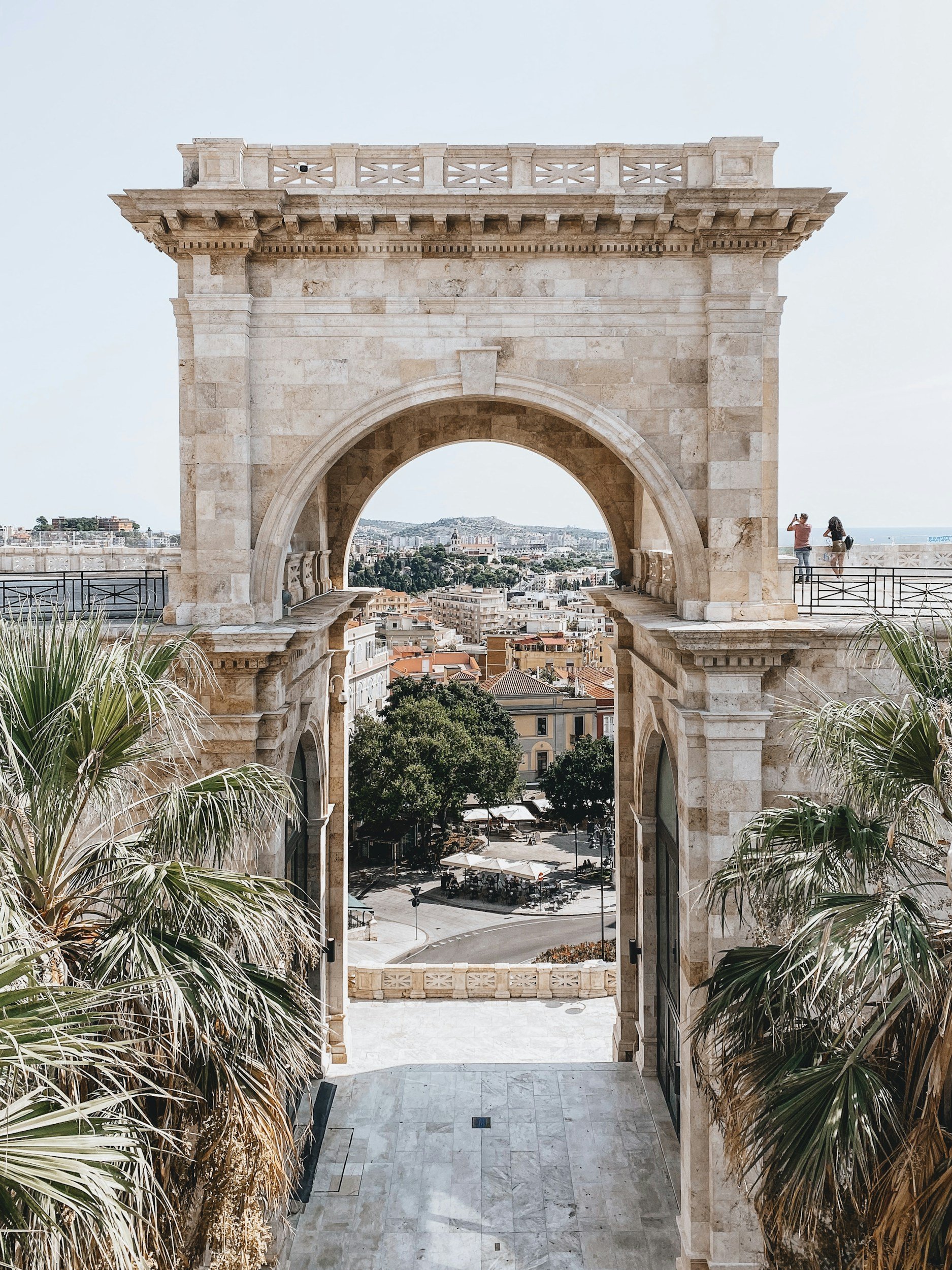 A historical stone archway overlooking a cityscape with buildings, trees, and a distant view of the sea, with blue sky overhead.