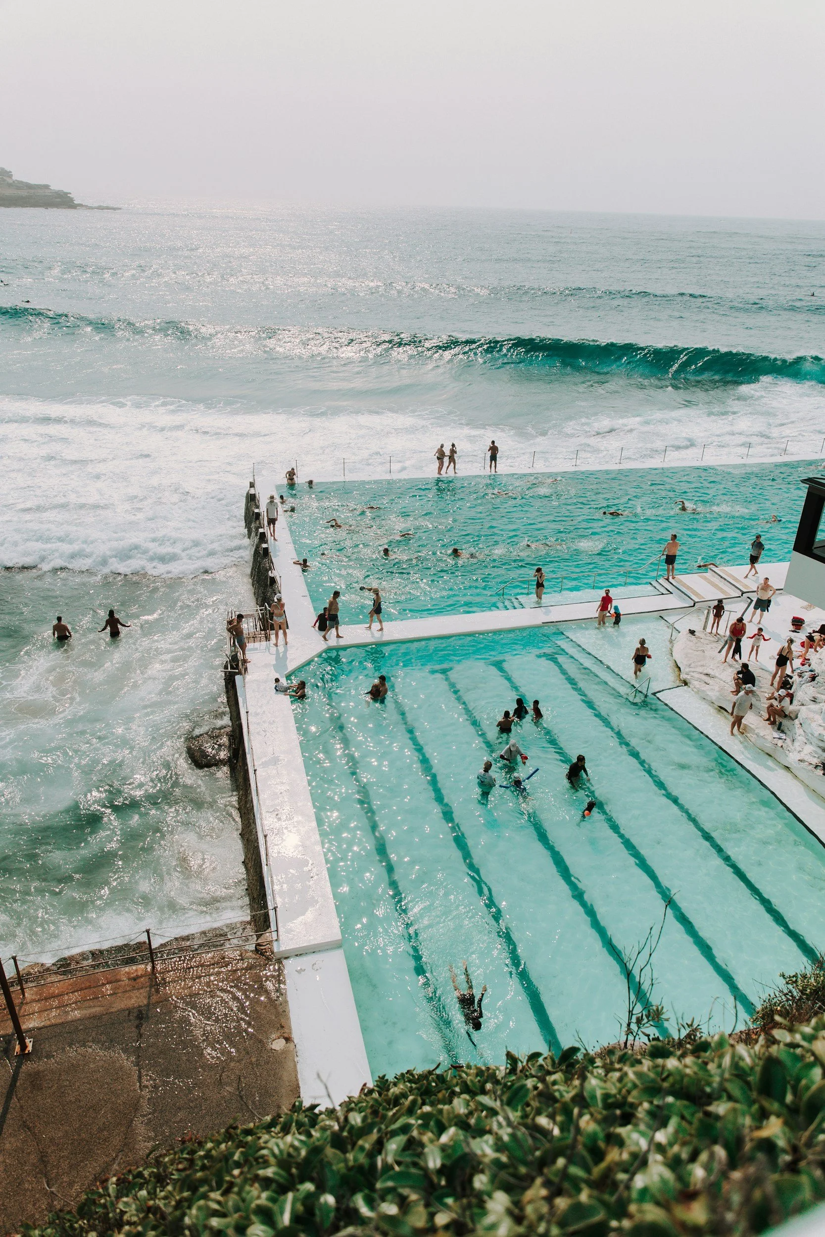 People swimming and relaxing in an outdoor pool near the ocean, with a view of waves and a foggy sky in the background.