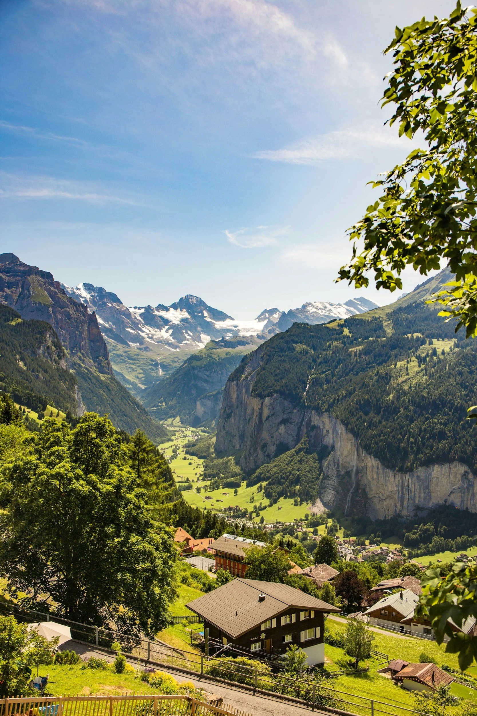 Scenic view of a lush green valley surrounded by tall mountains with snow-capped peaks under a blue sky with wispy clouds. There are houses and buildings in the foreground with trees and gardens.