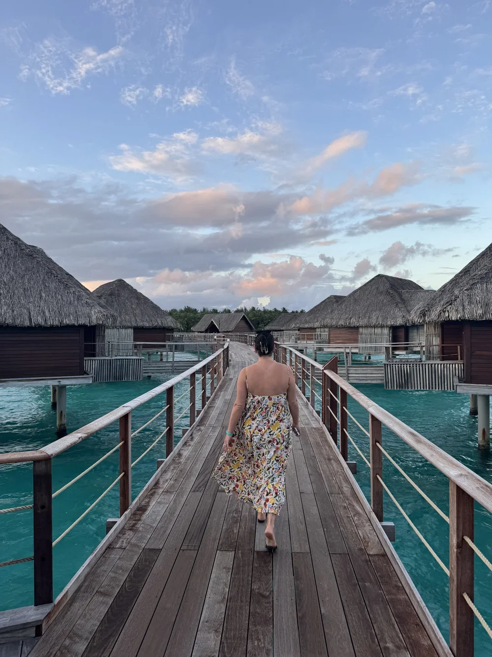 A woman in a floral dress walking on a wooden pier towards overwater bungalows under a sky with clouds.