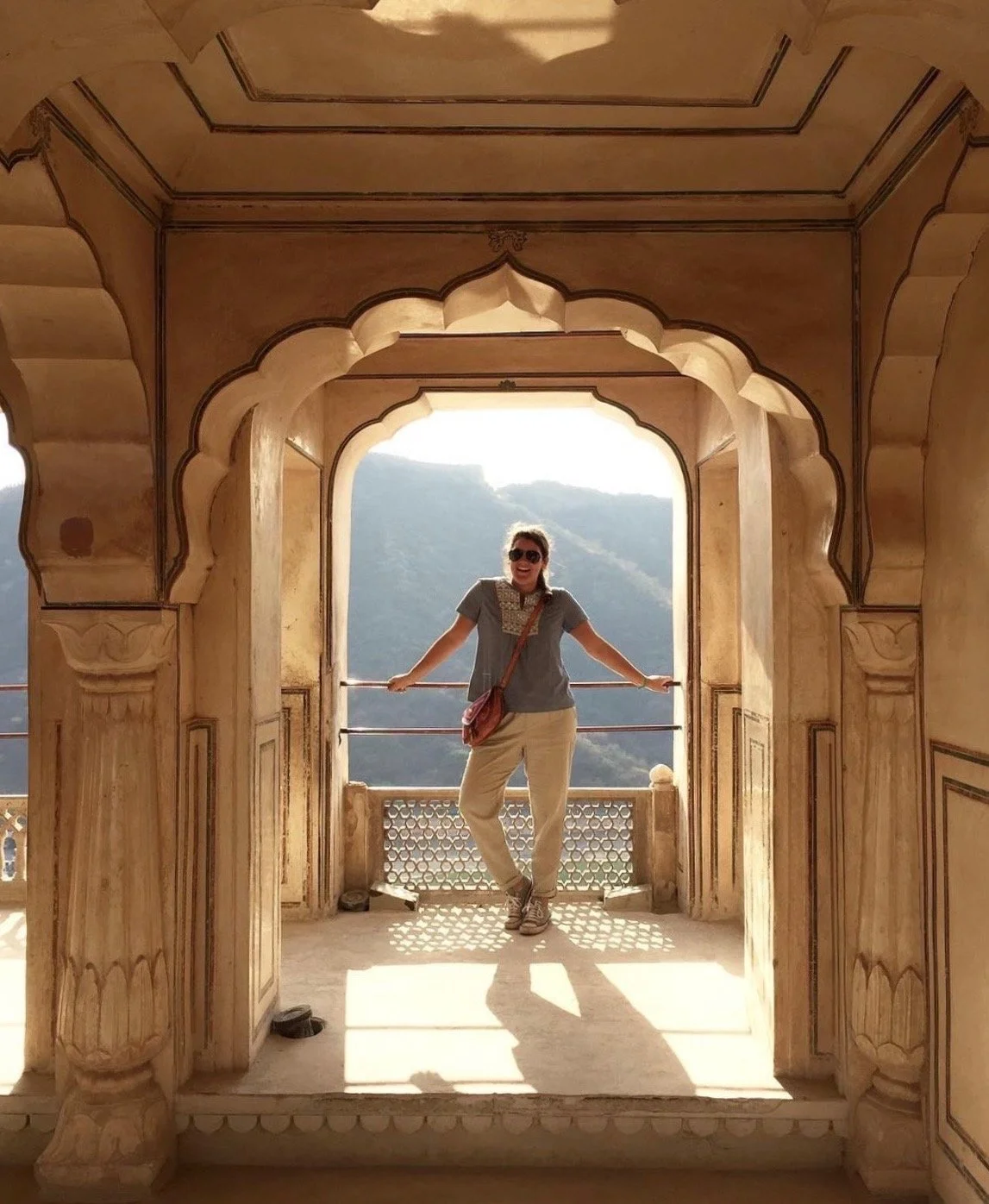 A woman standing in a carved stone archway with mountain scenery in the background, smiling and wearing sunglasses.