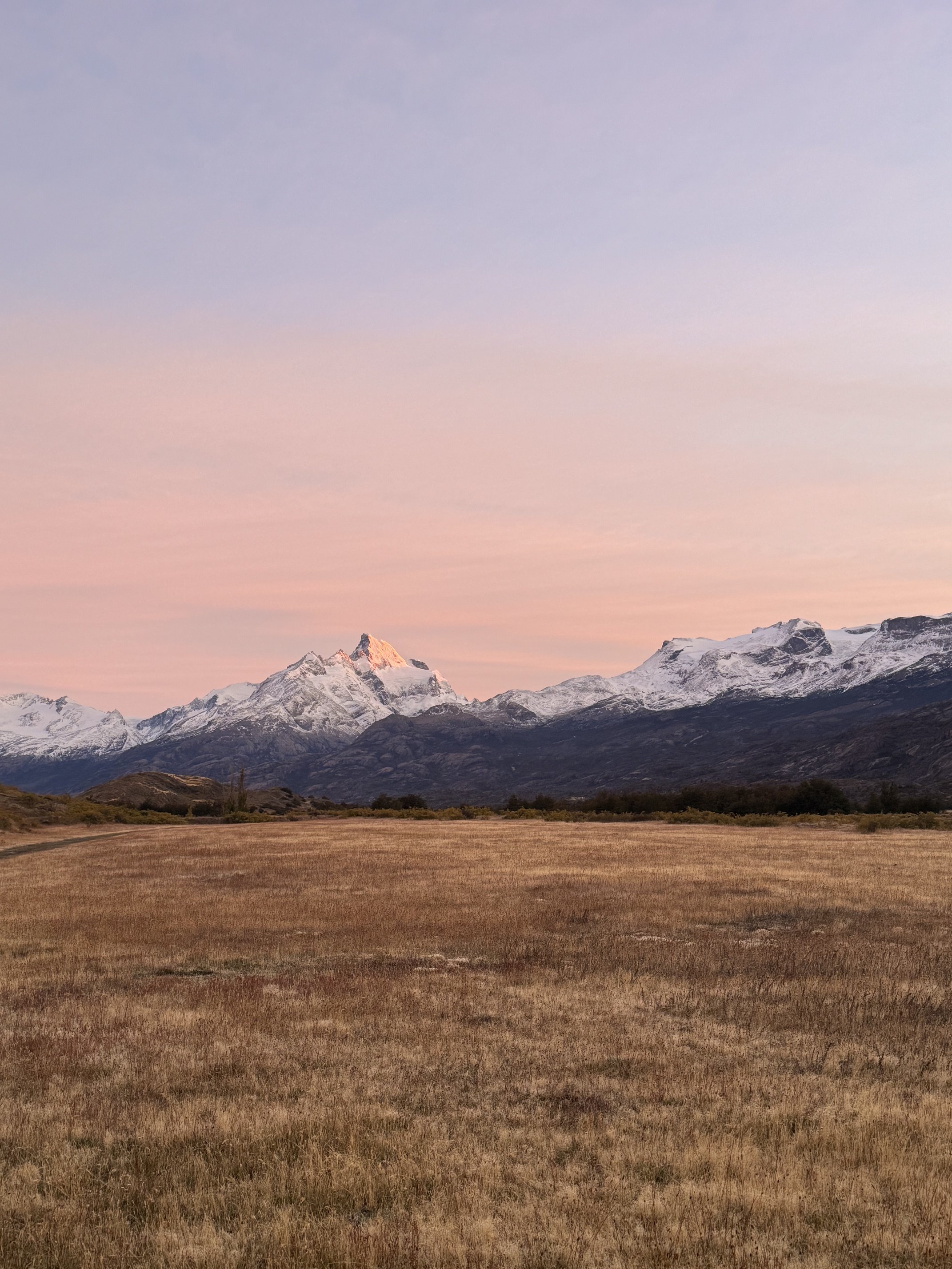 A mountain landscape at sunset with snow-capped peaks and a grassy field in the foreground.