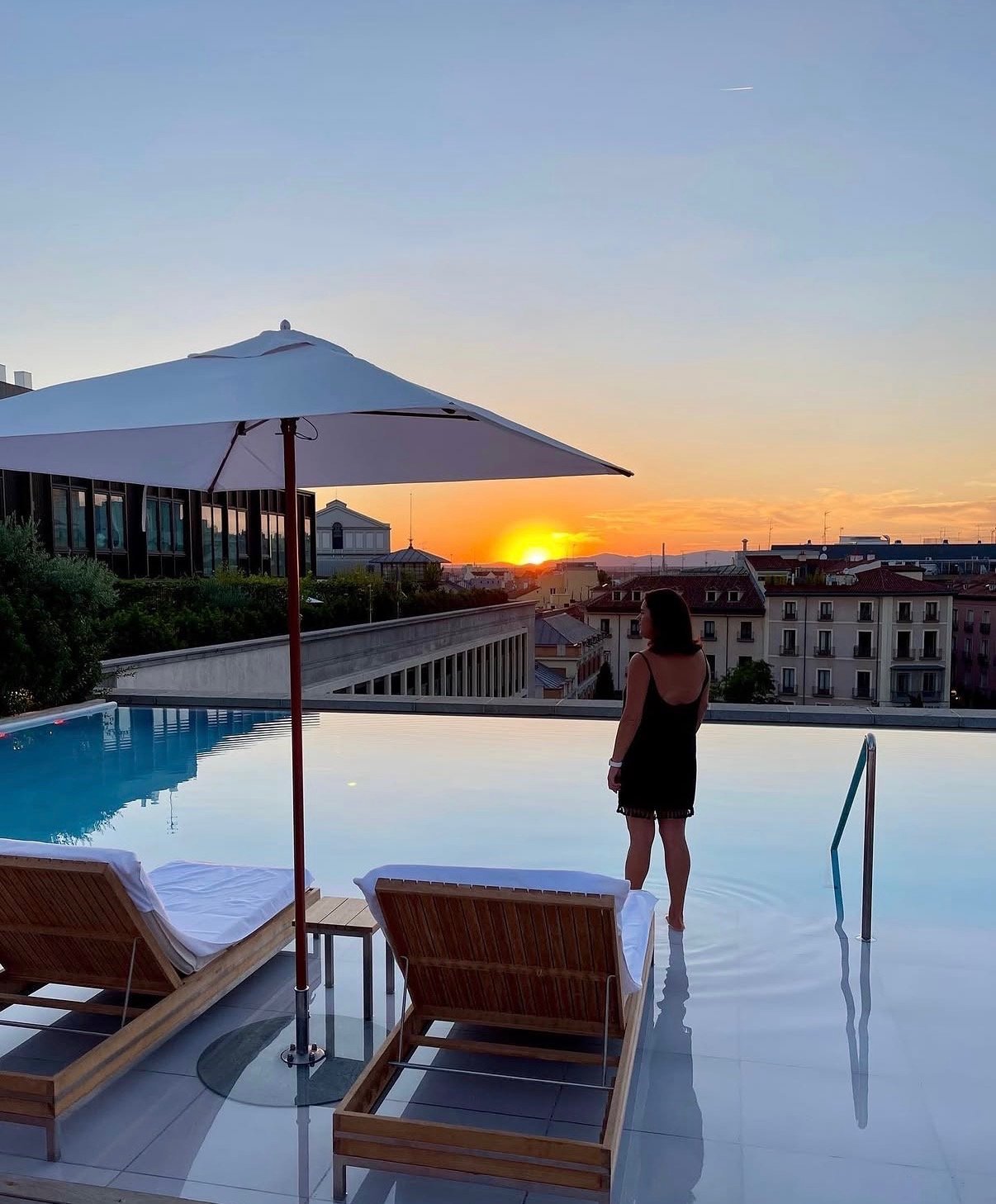 A woman standing at the edge of a rooftop pool during sunset, with an umbrella and lounge chairs nearby, overlooking a cityscape with buildings and mountains in the distance.