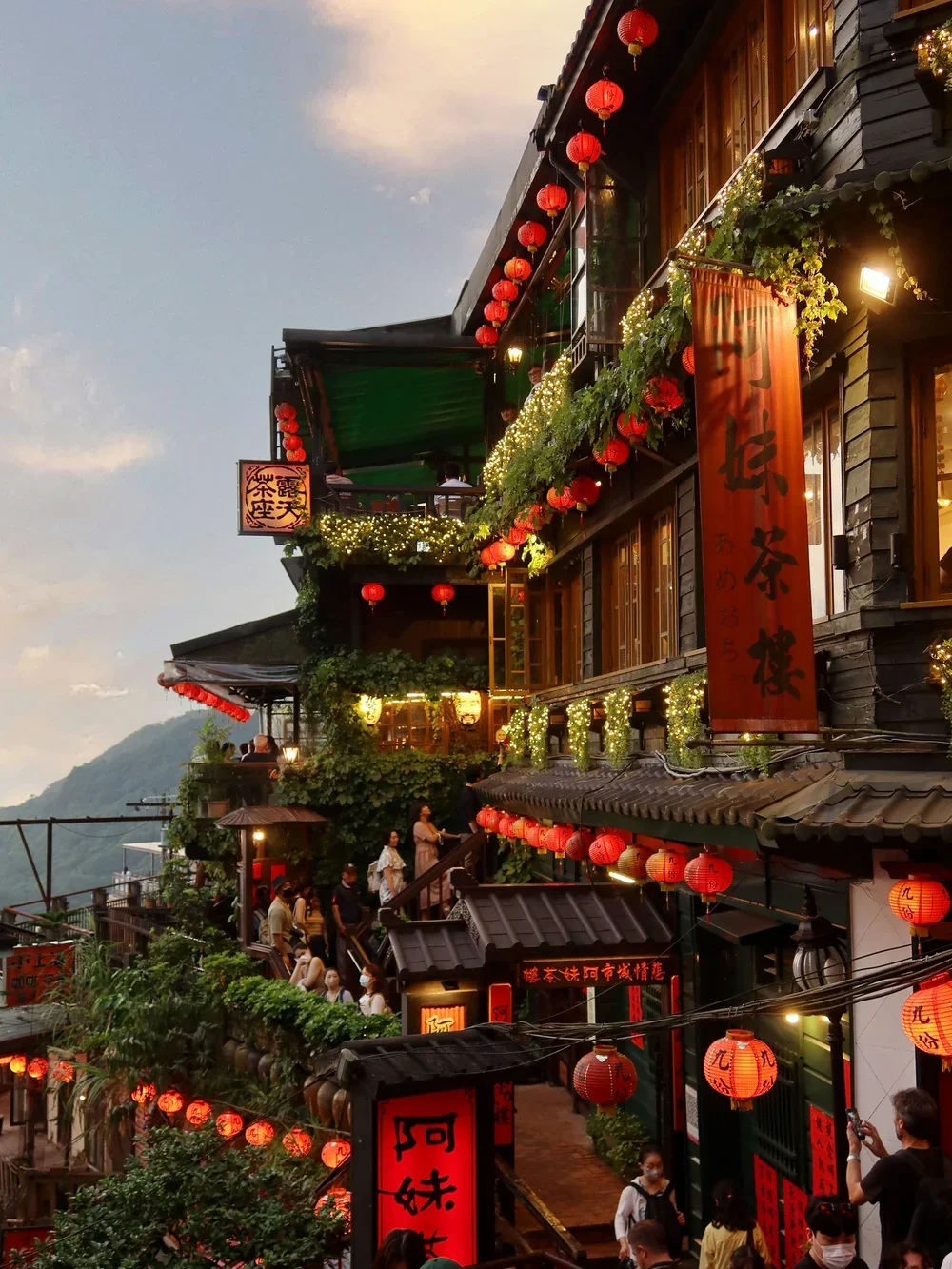 A multi-story building decorated with red lanterns, string lights, and greenery in an Asian style, with people dining and walking outside during dusk.