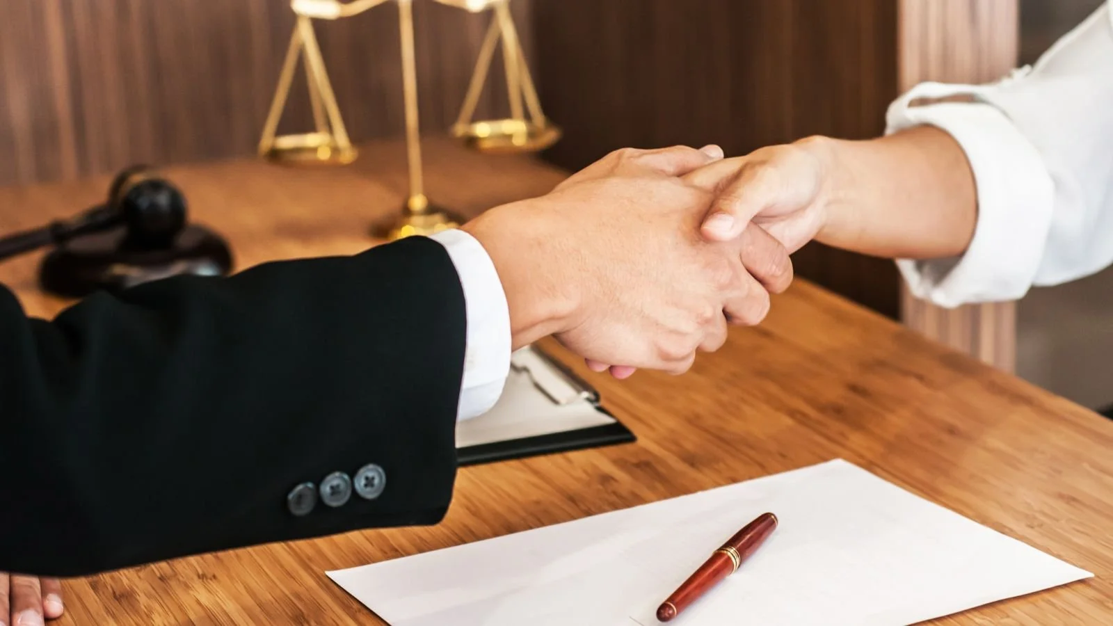 Person in a black suit shaking hands with person in a white shirt across a wooden desk. A pen and a paper are on the desk, with laboratory scales in the background.