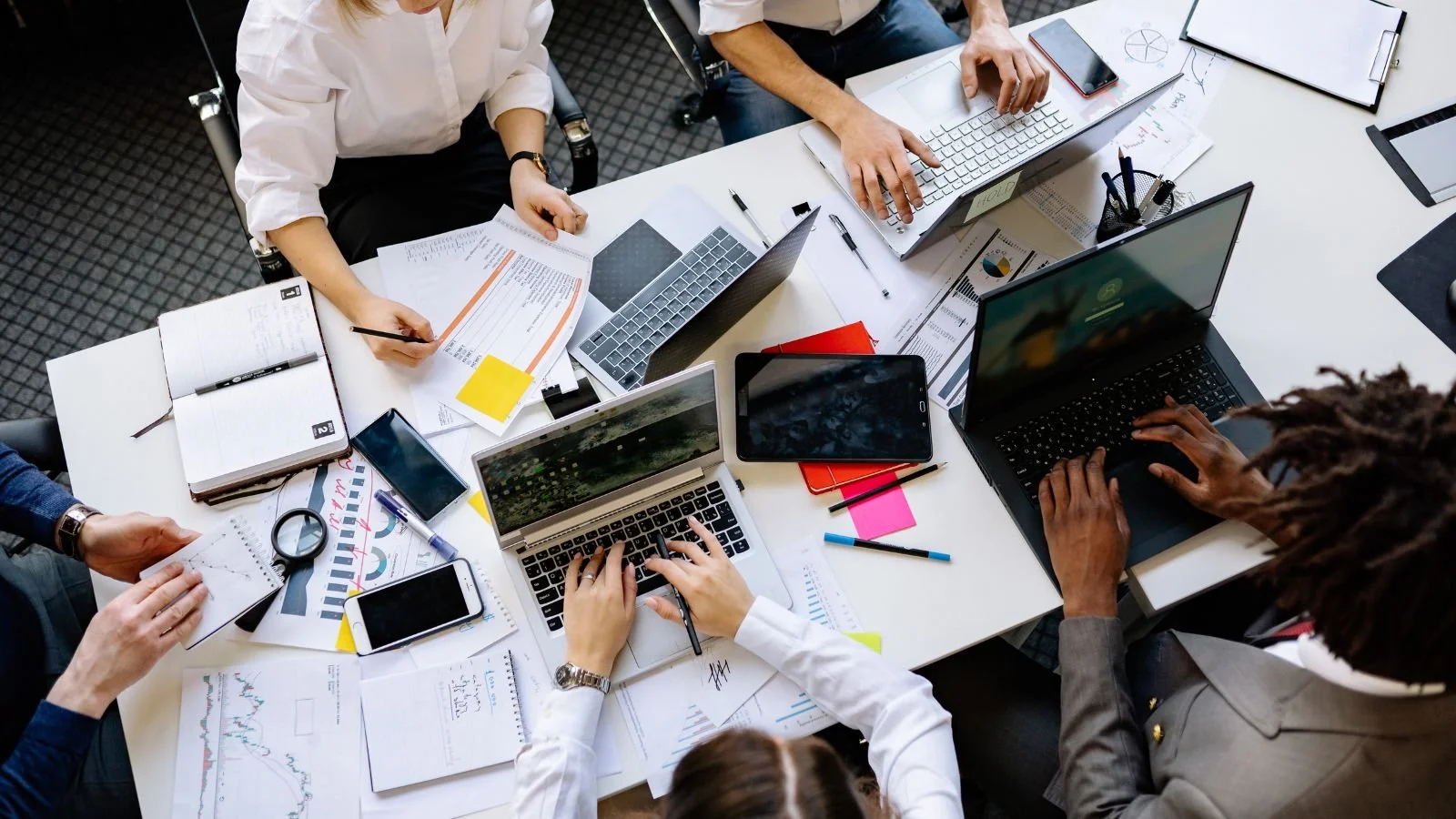 Top-down view of a busy office meeting table with five people working on laptops, with papers, notebooks, pens, and smartphones scattered on the table.