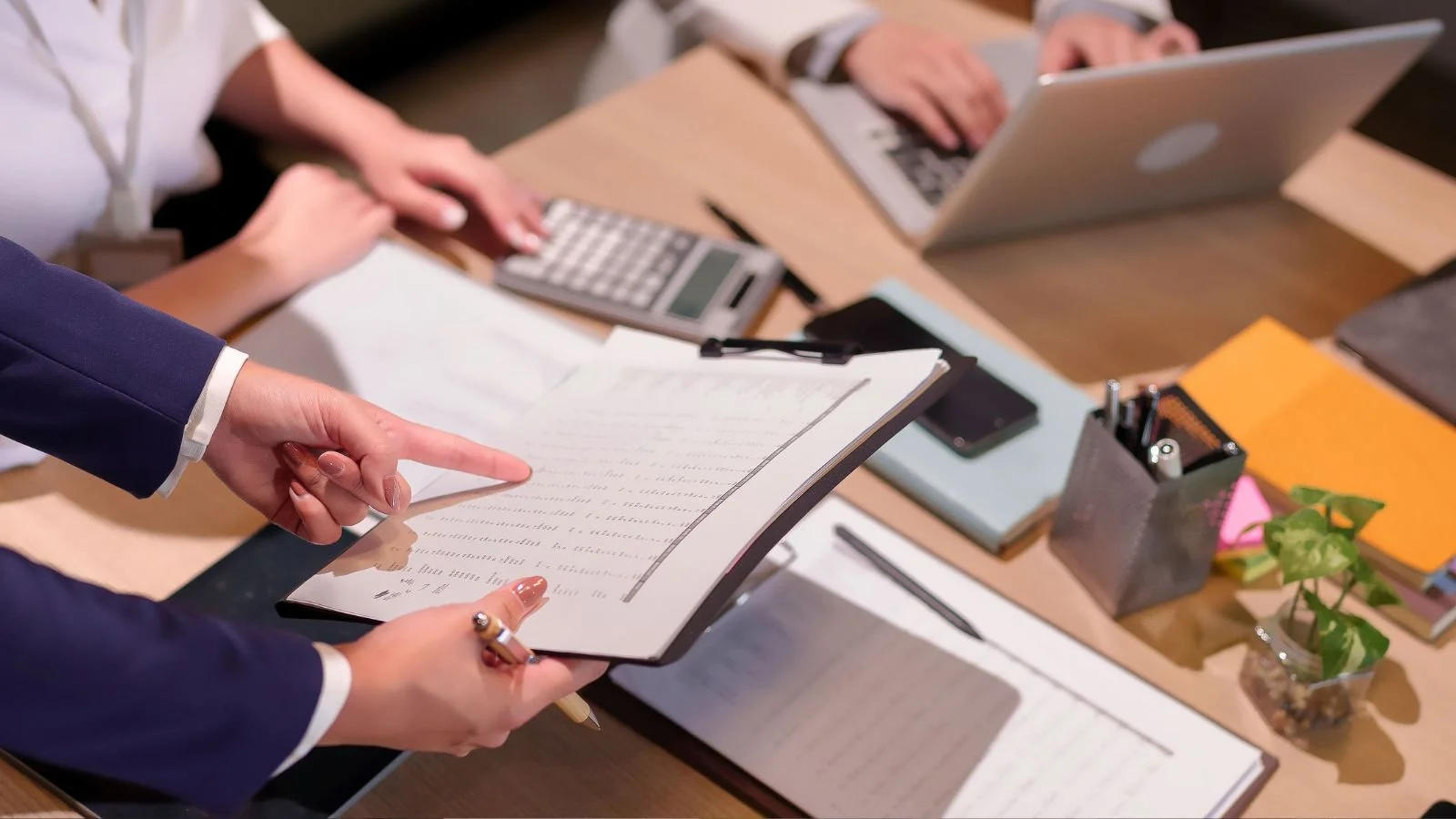 People working at a desk with laptops, notebooks, a calculator, pens, and a potted plant.