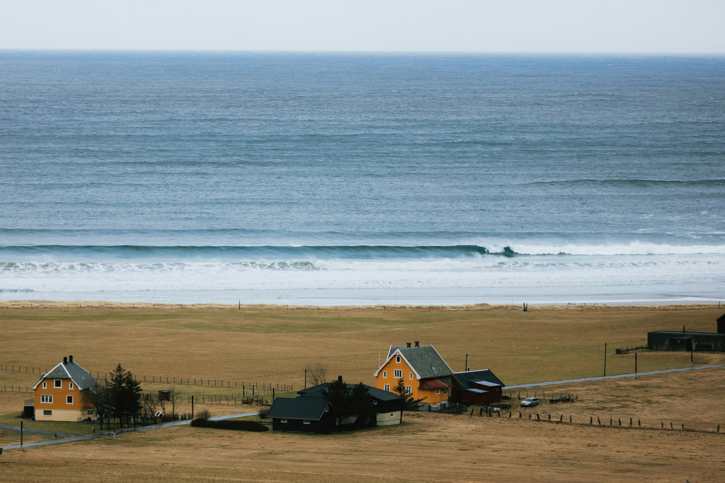 A scenic view of the ocean with waves reaching the shoreline, and a small residential area with houses at the edge of a grassy field.