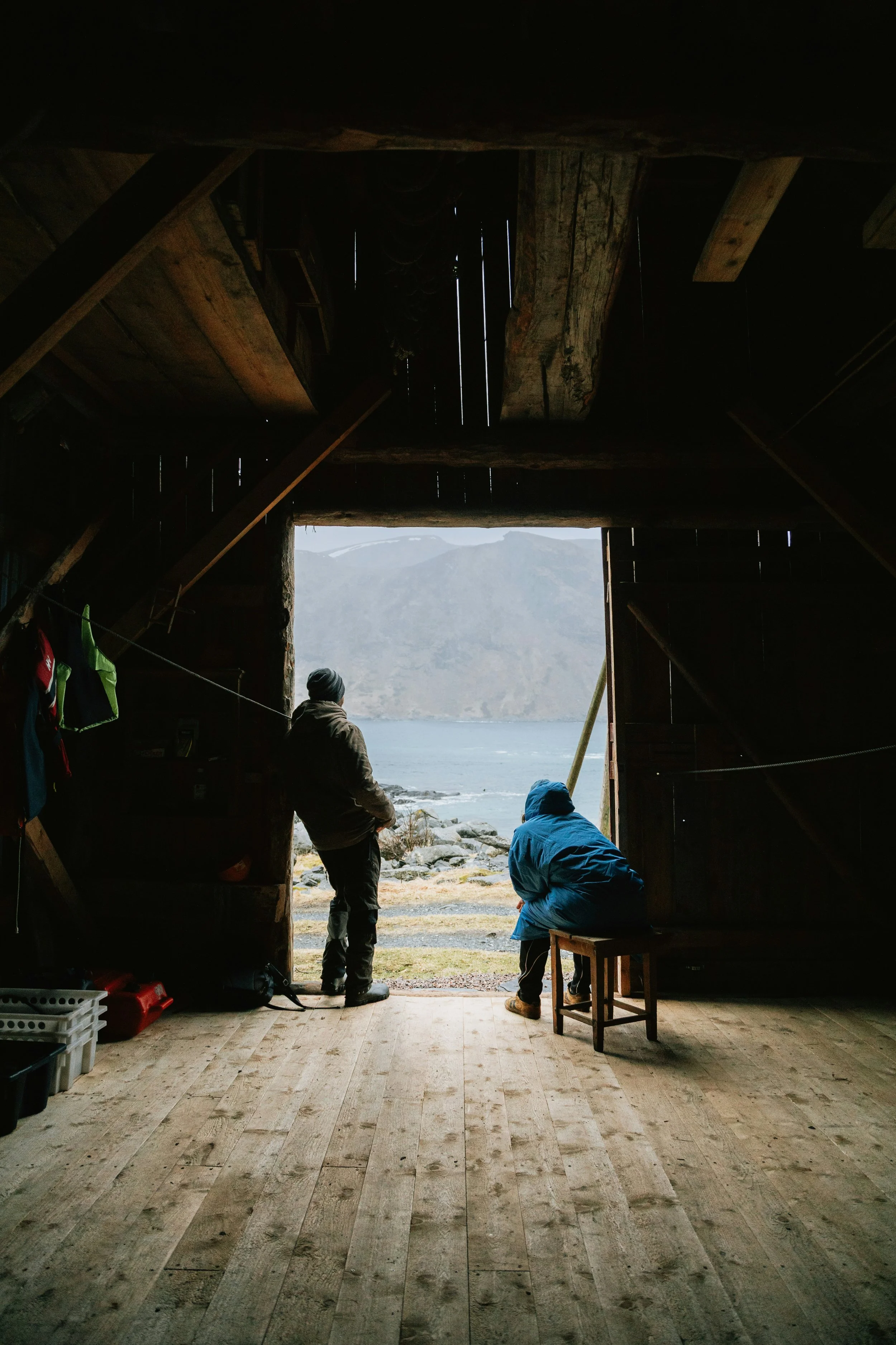 View from inside a rustic wooden cabin or shed, looking out towards a body of water with mountains in the background. Inside, there are two people, one standing and one sitting, dressed in outdoor gear, possibly preparing for outdoor activities like fishing or hiking. The interior has a wooden floor and various gear hanging or stored on the sides.