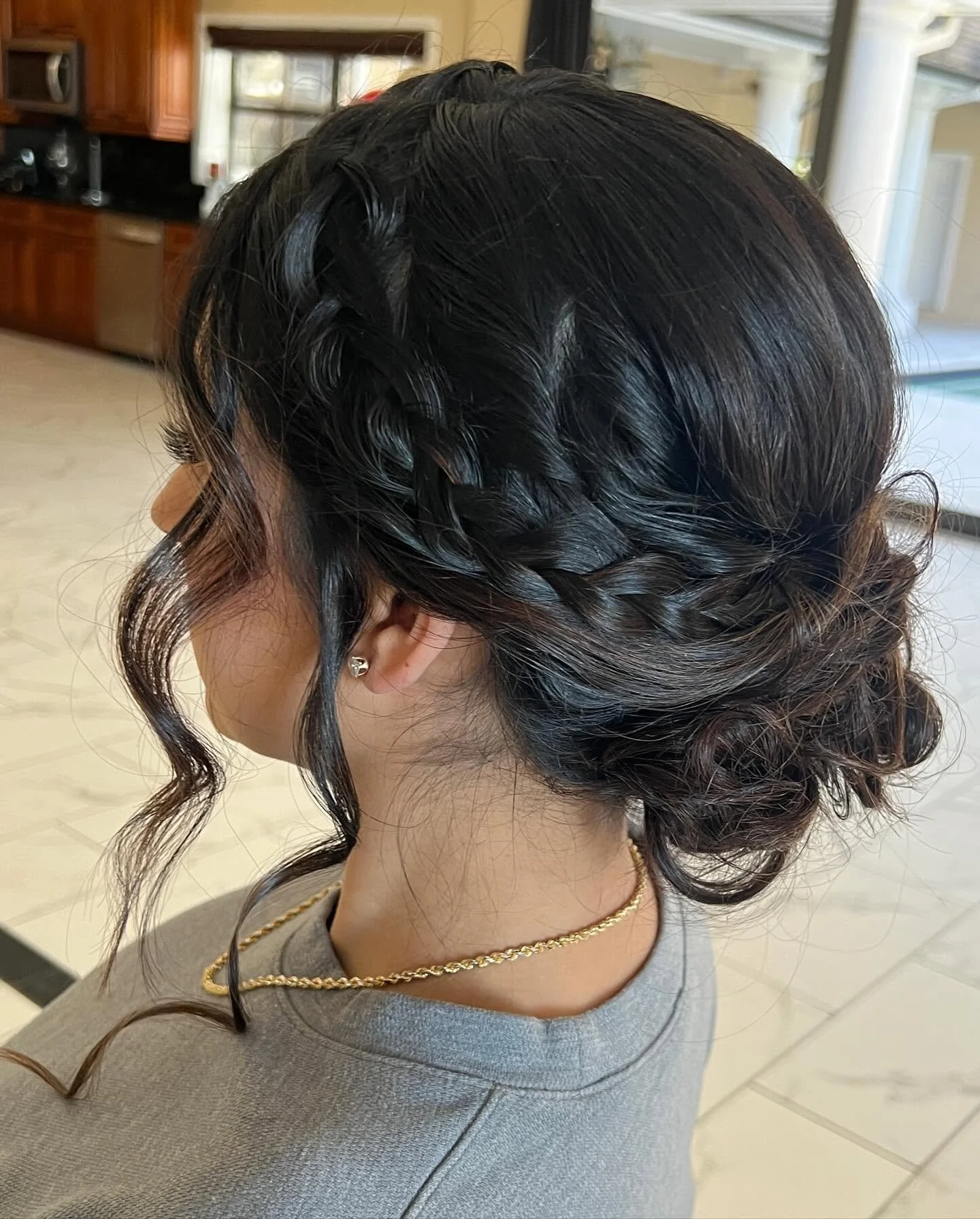 A woman with dark hair styled in an elegant updo with intricate braids and curls, wearing small earrings and a gold necklace, sitting in a room with large windows and wooden cabinets.