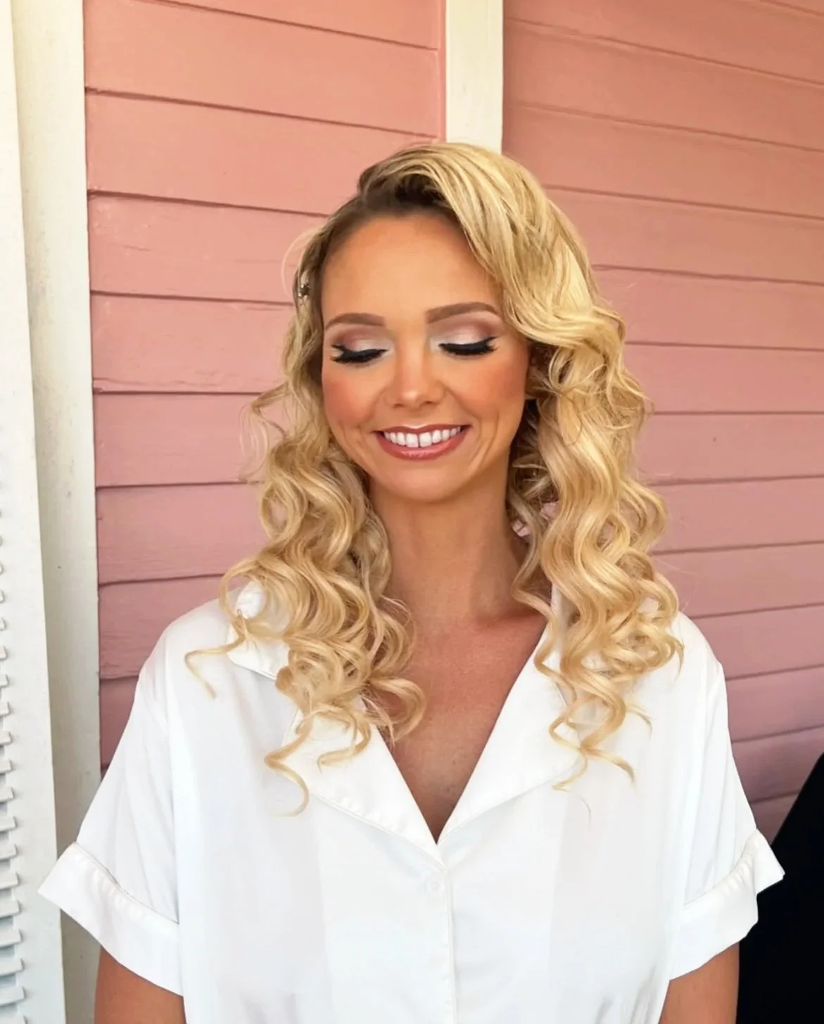 A woman with blonde, curly hair smiling with closed eyes, wearing makeup and a white shirt, standing in front of a pink wooden wall.