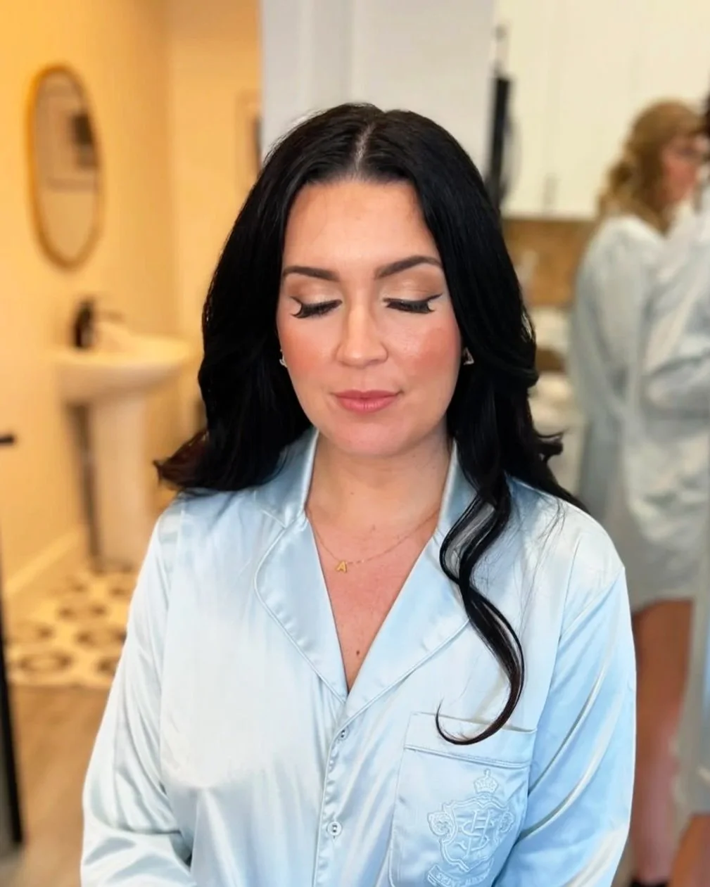 A woman with long black hair and light makeup, wearing light blue satin pajamas, standing in a room with a warm yellow wall, a mirror, a sink, and a person in medical clothing in the background.
