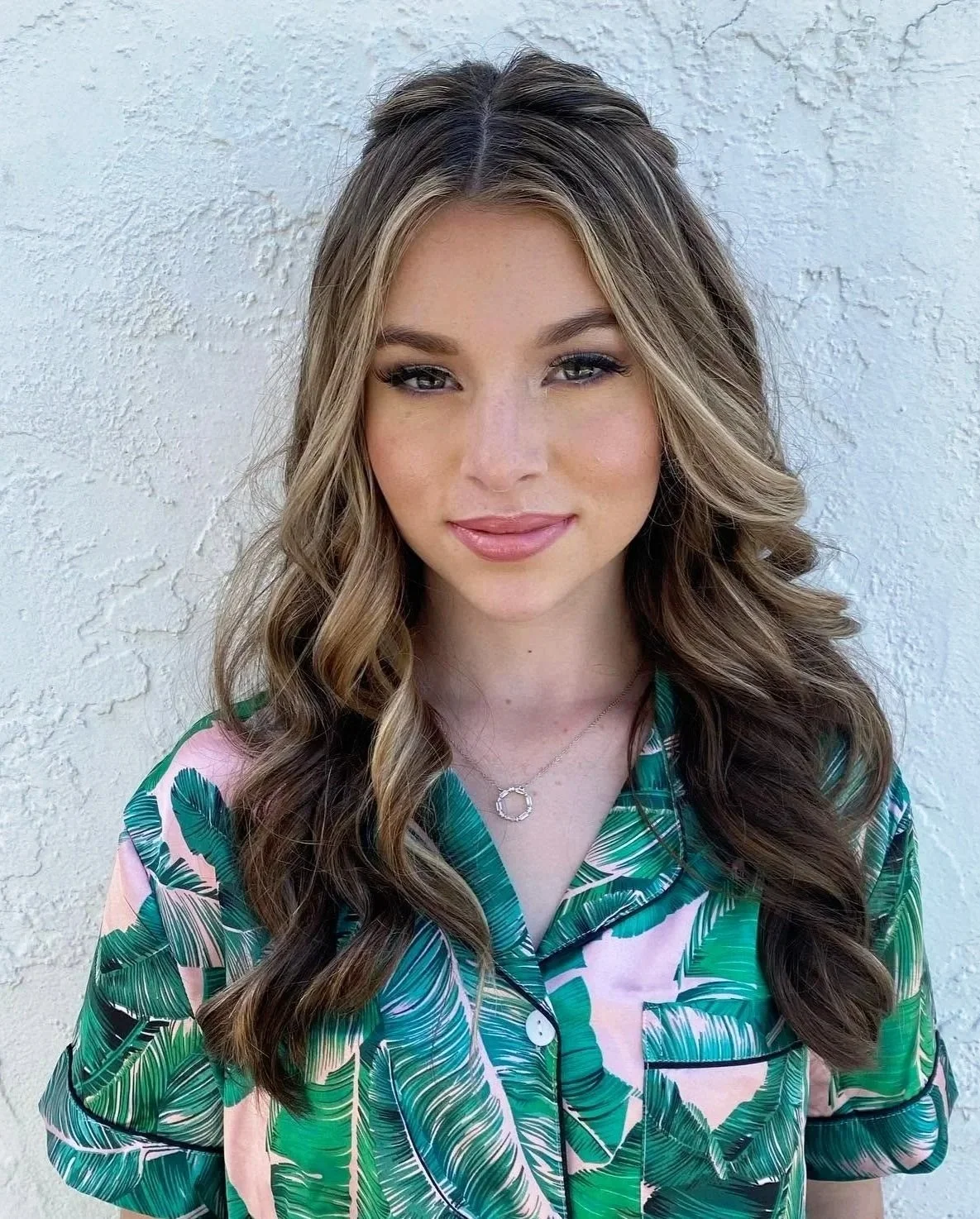 A young woman with long, wavy brown hair and light makeup standing against a white textured wall. She is wearing a silk button-up shirt with a green and pink tropical leaf pattern and a silver necklace with a circular pendant.