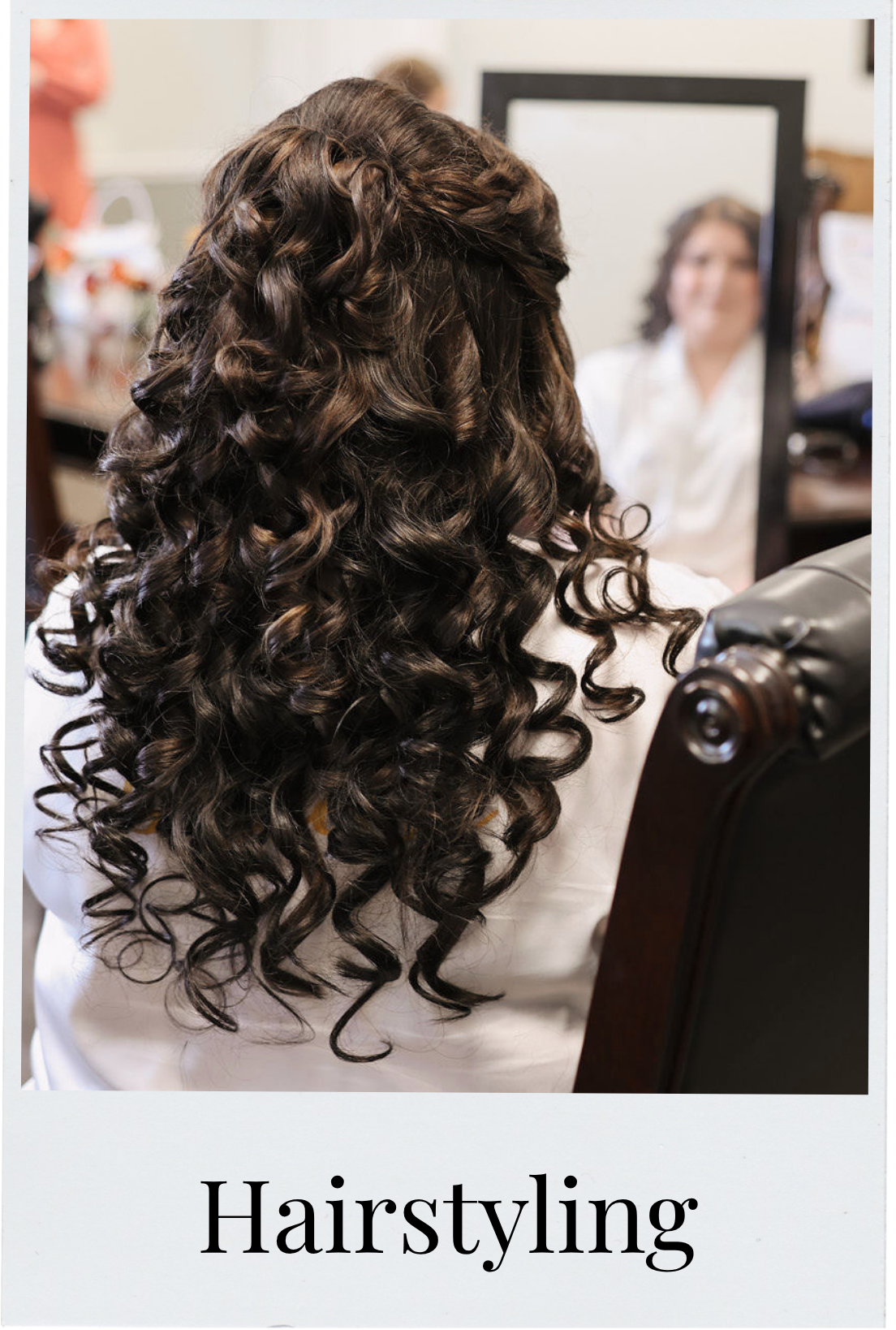 A woman with long, dark, curly hair sitting in a salon chair, looking at herself in a mirror, during hairstyling.