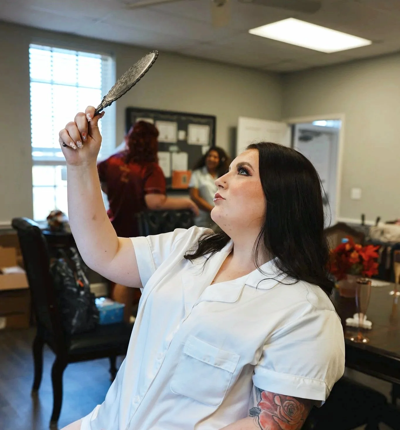 Woman with black hair and makeup holding a decorative mirror in a room with other women and decorated with flowers.