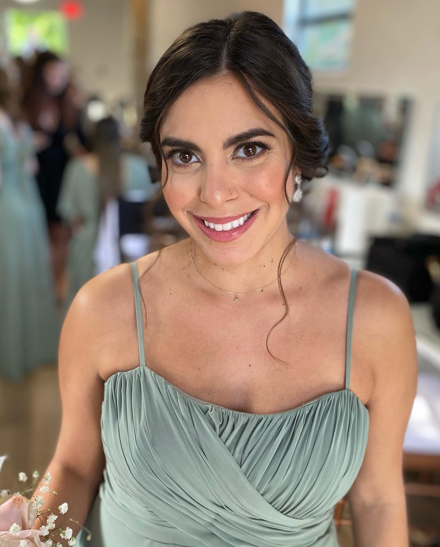 A smiling woman with dark hair styled in an updo, wearing a light green dress with spaghetti straps and a delicate necklace with an 'A' pendant. She is indoors at a social event, with blurred people and decor in the background.