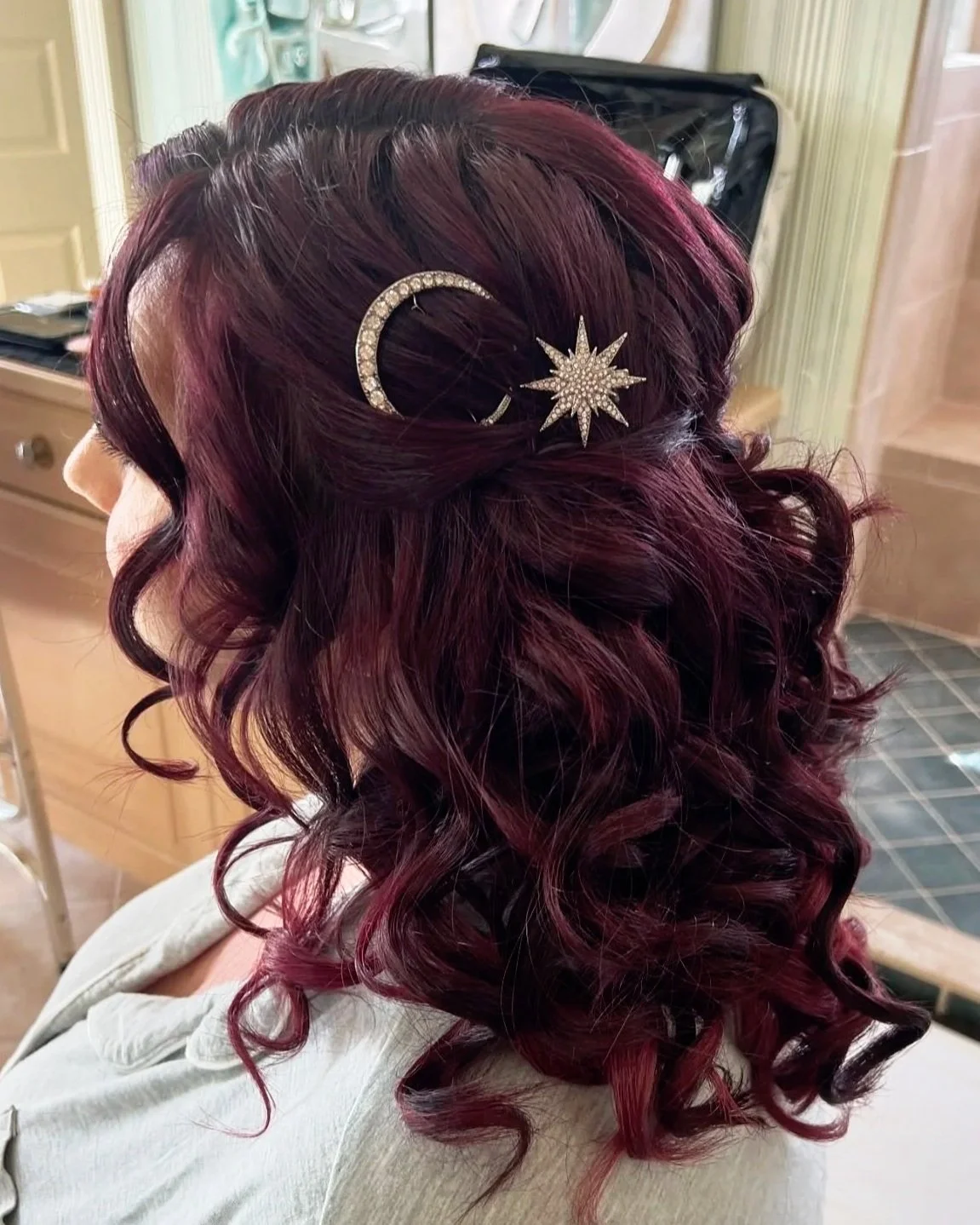 A woman's hairstyle with dark red, curly hair decorated with a crescent moon and star-shaped rhinestone hair clips.