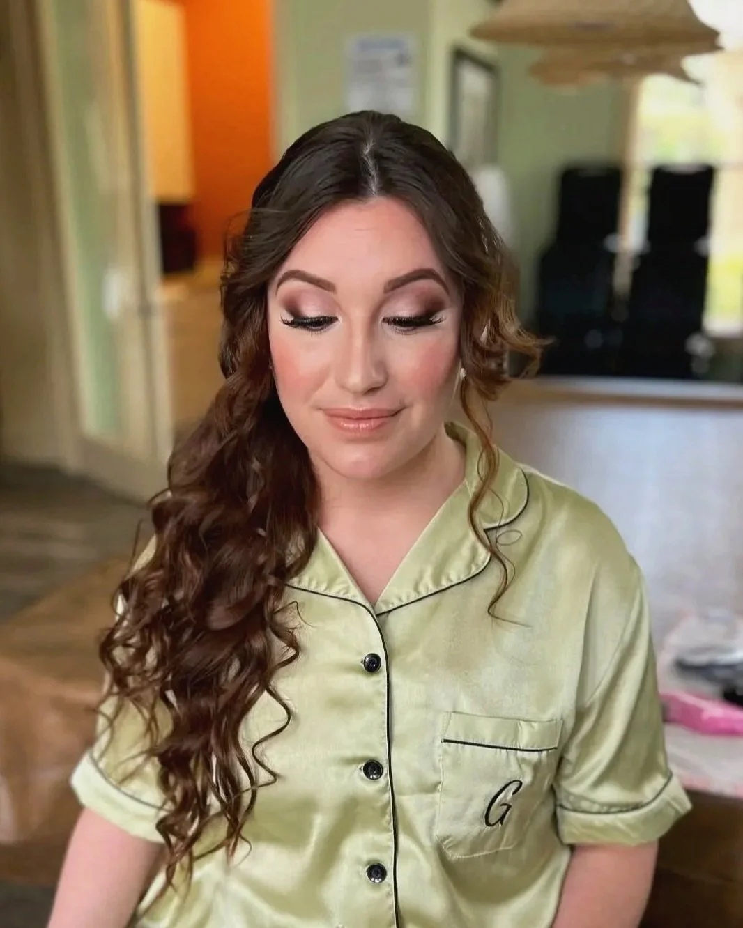 A woman with long, curly brown hair and makeup, wearing a yellow satin pajama top with black piping and the letter G embroidered on it, smiling with eyes closed.