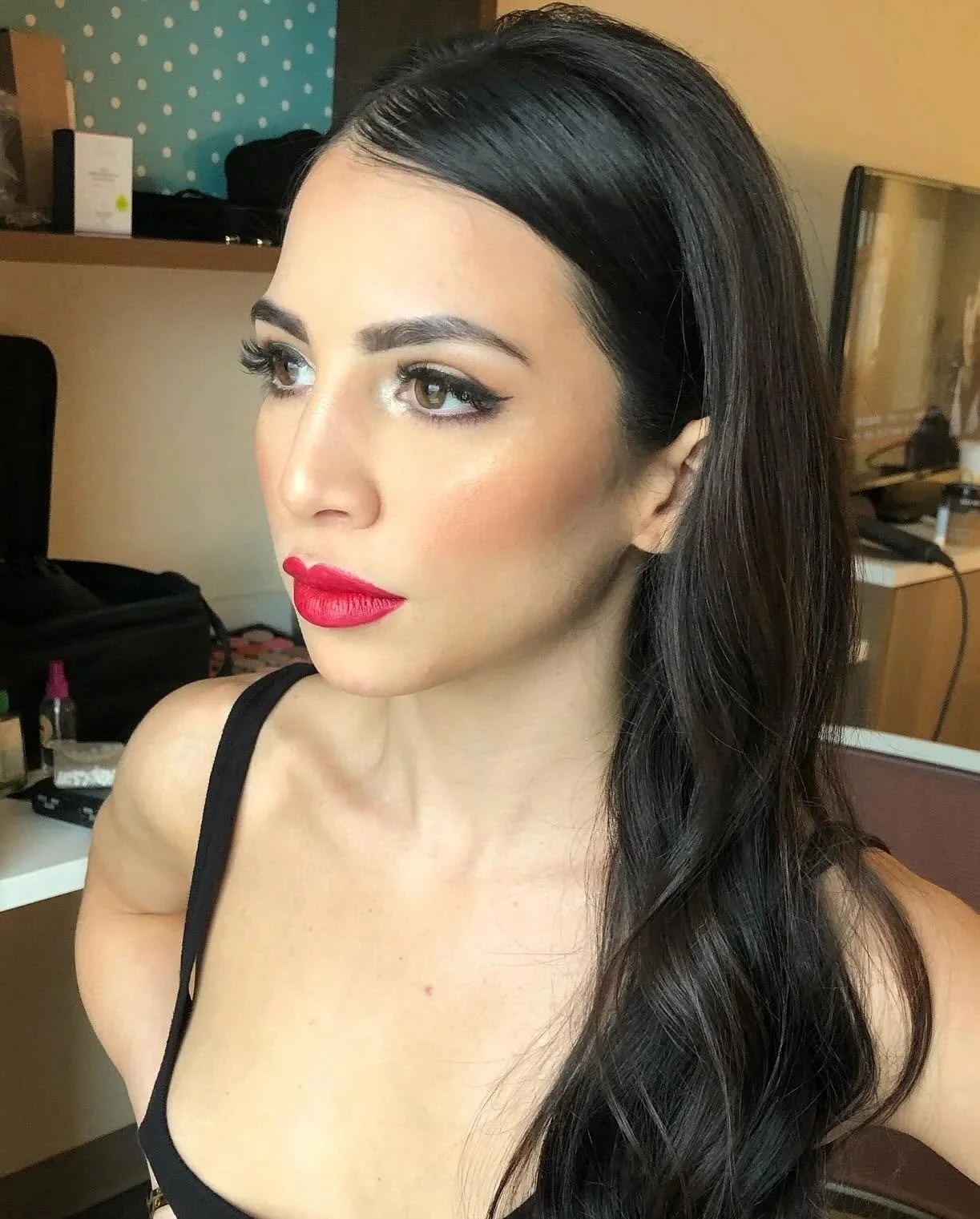 Close-up of a woman with long dark hair, makeup, red lipstick, wearing a black top, in a room with makeup and beauty tools in the background.