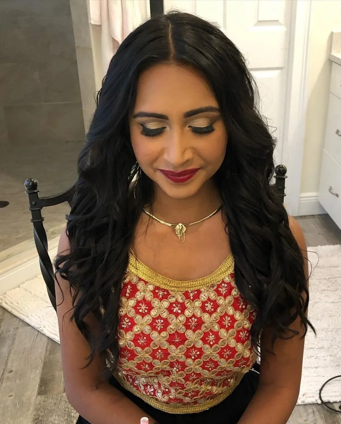 A woman with long black wavy hair, closed eyes, and red lipstick, wearing a red and gold embroidered top, a gold necklace, and sitting on a chair in a room with a door and a white dresser in the background.