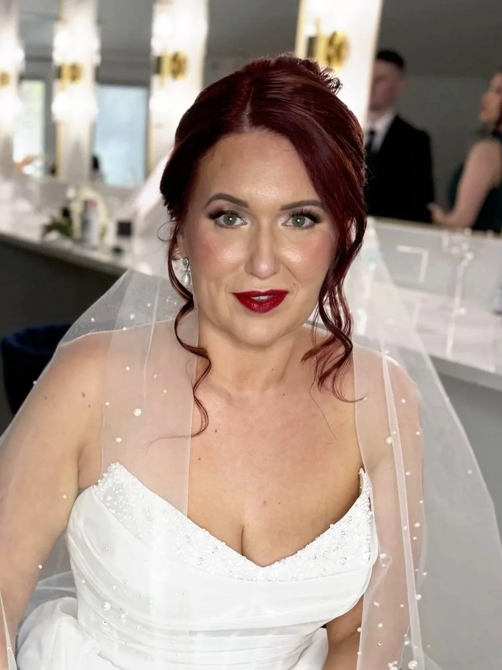 A woman with red hair and fair skin dressed in a white wedding gown with a tulle veil, sitting in a backstage area with mirrors and other people in the background.