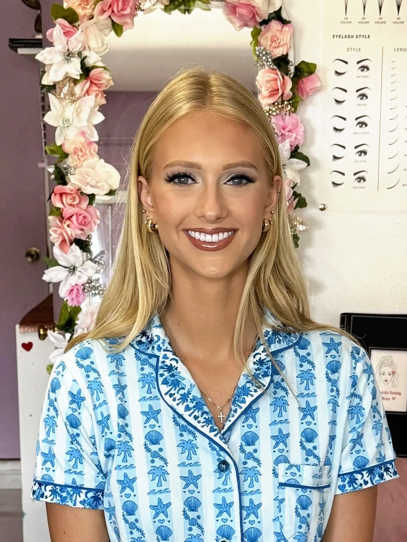 A woman with blonde hair and blue eyes smiling, wearing blue and white patterned pajamas with starfish and seashells, standing in front of a floral wreath and a chart about eyelash styles.