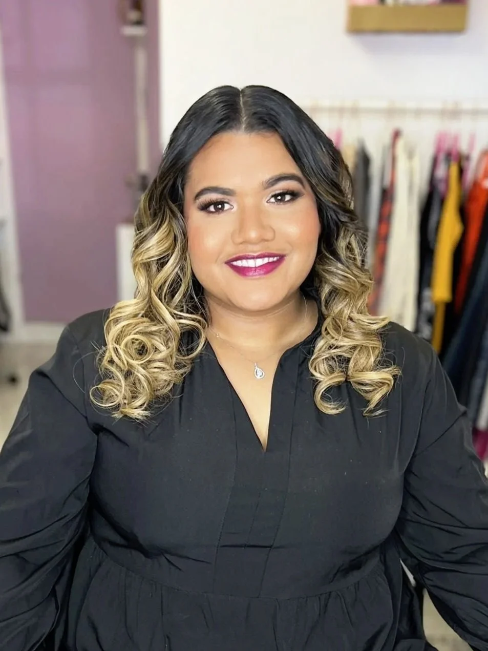 A woman with curly, ombre hair smiling, wearing makeup and a black top, standing in a room with a clothing rack in the background.
