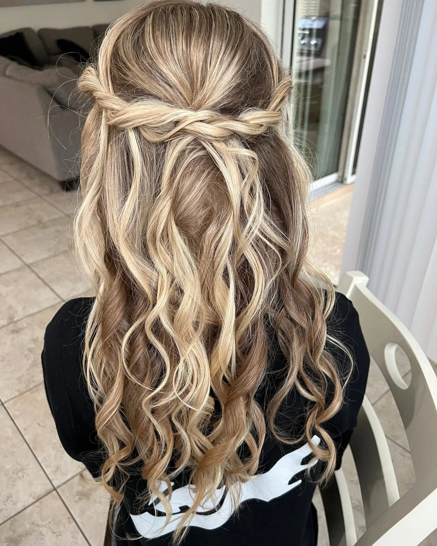 A person with long, wavy blonde hair styled with a twisted half-updo, sitting on a white chair in a room with tiled flooring and a sliding glass door.