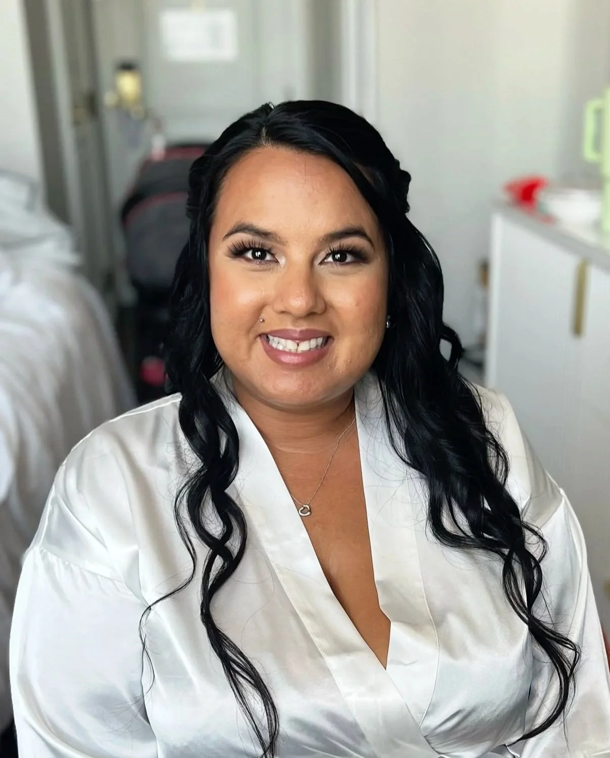 Smiling woman with long black hair, wearing a white satin top and a delicate necklace, sitting in a bright room with blurred background.