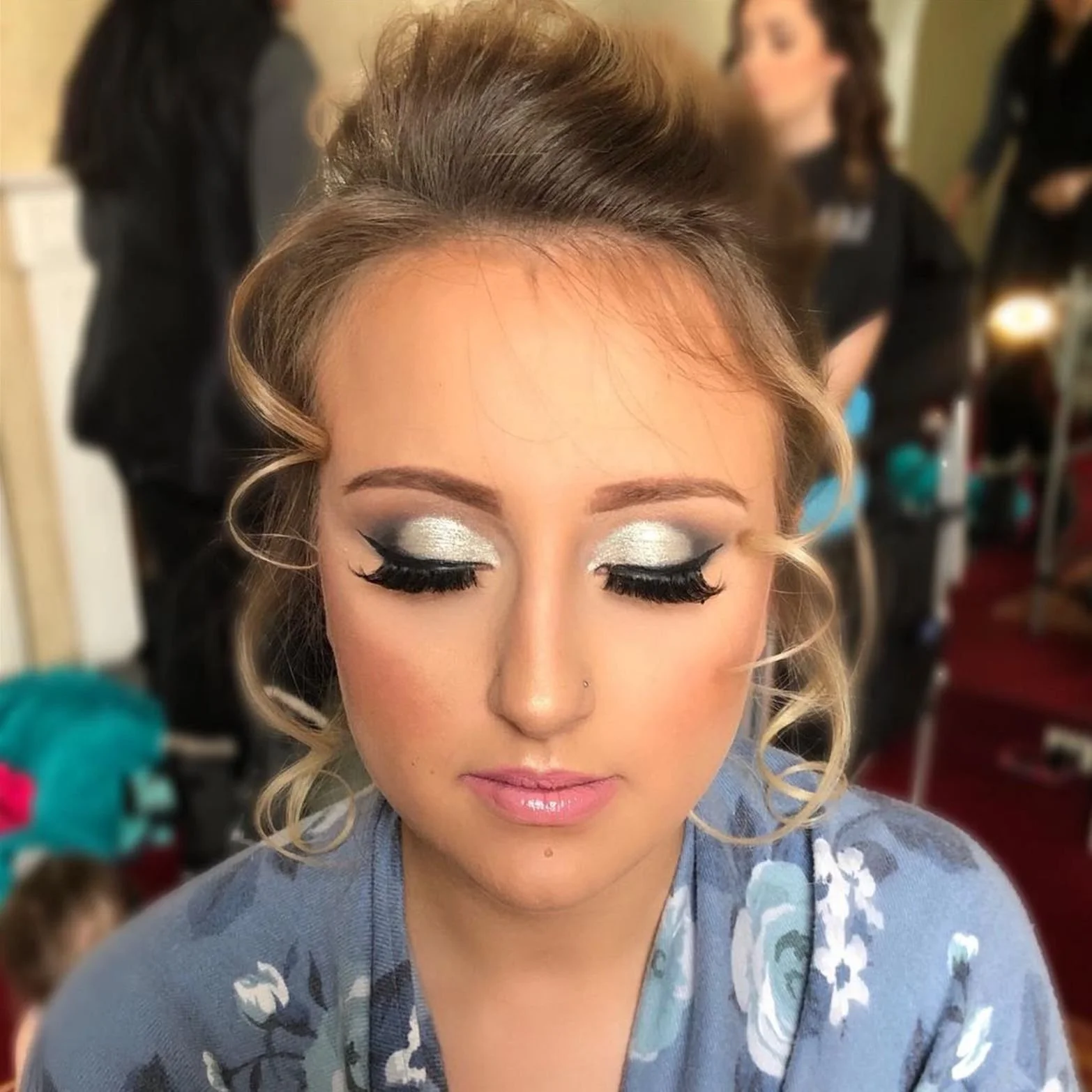 Close-up of a woman with styled curly blonde hair and makeup, including silver eyeshadow and black eyeliner, with other women in the blurred background at a makeup or fashion event.