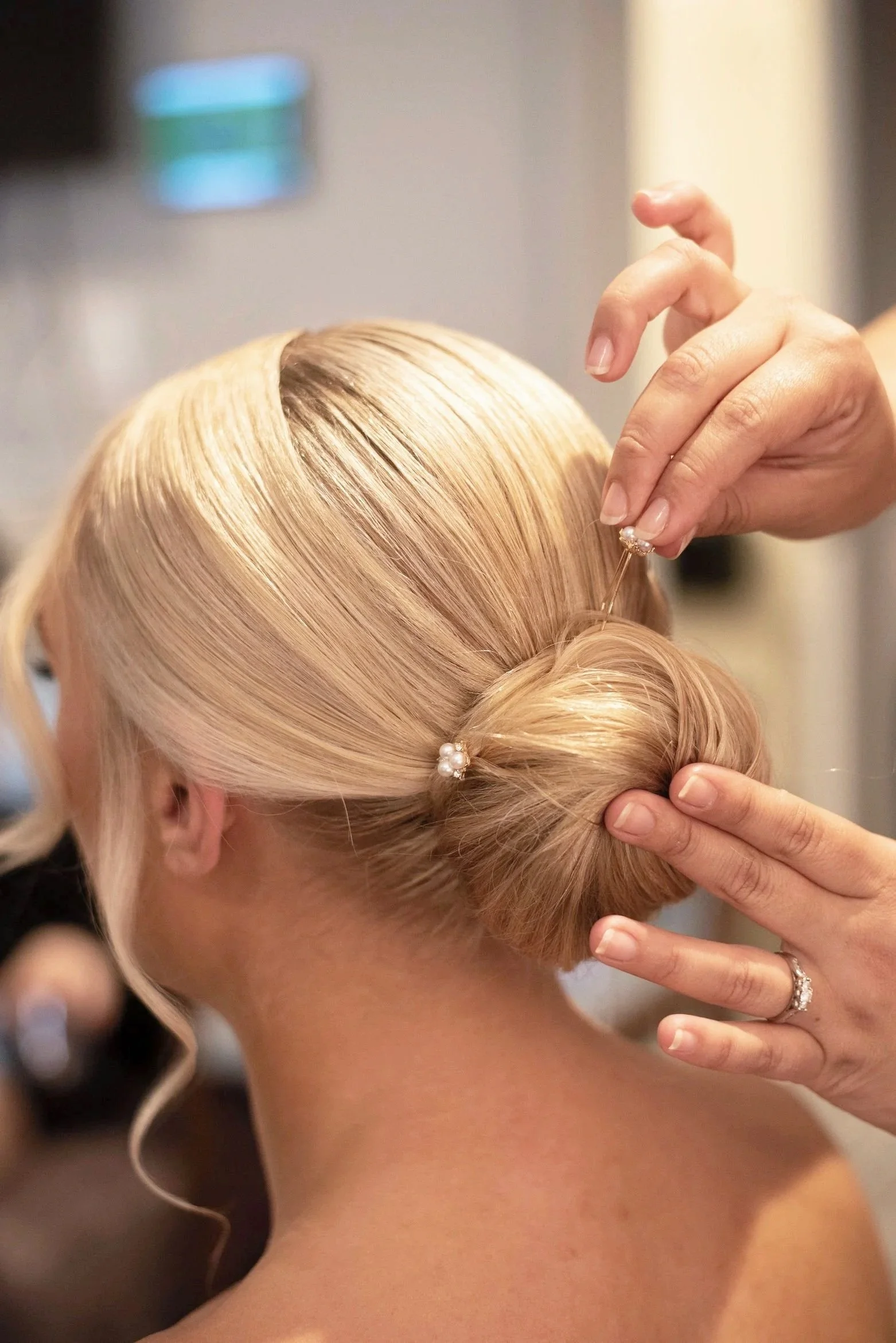 A person with blonde hair styled in an elegant updo, wearing pearl hair accessories, is having their hair styled by a hairstylist who is adding a decorative hairpin.