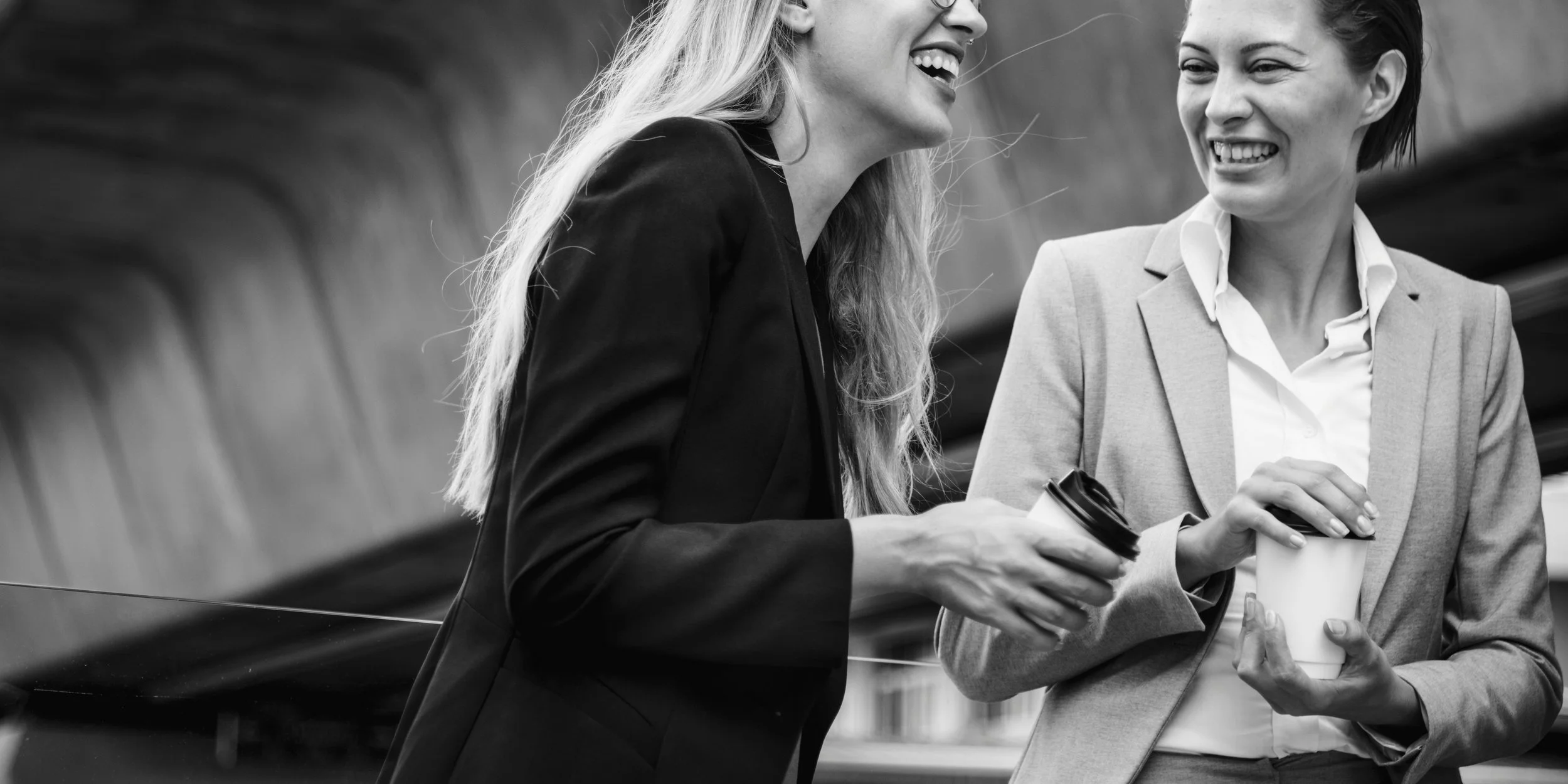Two women laughing and conversing over a cup of coffee, dressed in corporate clothing