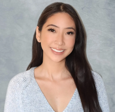 A young woman with long dark hair smiling at the camera, wearing a light-colored top, against a plain gray background.