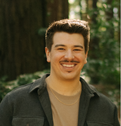 Smiling man (Wyatt Ousey) with dark hair, mustache, wearing a black jacket over a beige shirt, standing outdoors in a wooded area.