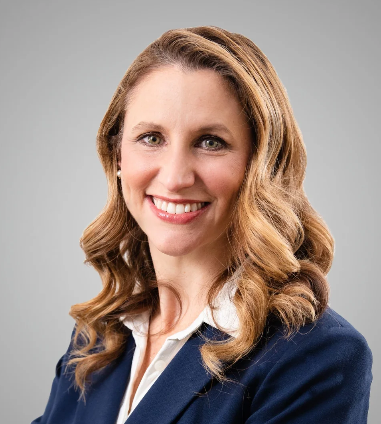 Professional woman (Danelle Van Metre) with wavy blonde hair wearing a navy blazer and white shirt, smiling against a gray background.