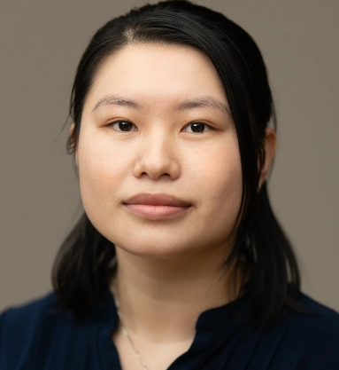 A woman with black hair wearing a dark blue top, looking directly at the camera against a neutral background.