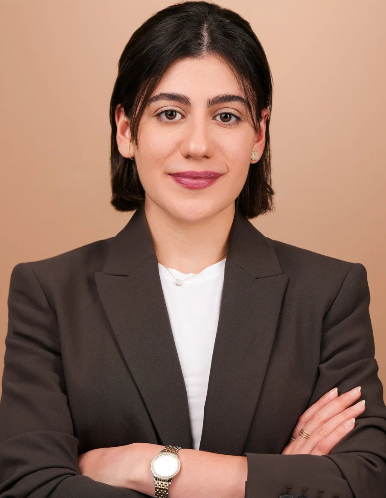 Professional woman (Amani Ezzo) with short dark hair, wearing a dark blazer and white shirt, with arms crossed, standing against a neutral background.