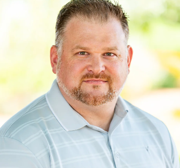 A man with short hair, a beard, and mustache, wearing a light blue polo shirt, smiling slightly against a blurred outdoor background.