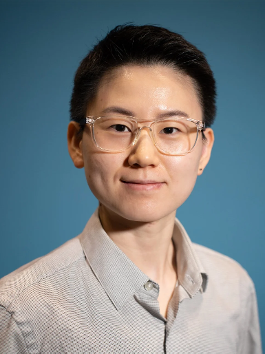 Portrait of a person with short dark hair, wearing glasses and a light-colored collared shirt, smiling against a blue background.