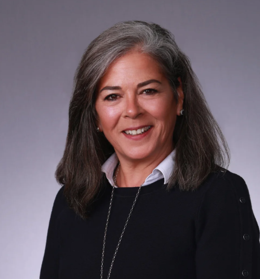 Smiling woman (Suzanne Ousey) with shoulder-length gray hair, wearing a white collared shirt, black sweater, and a necklace, against a gray background.