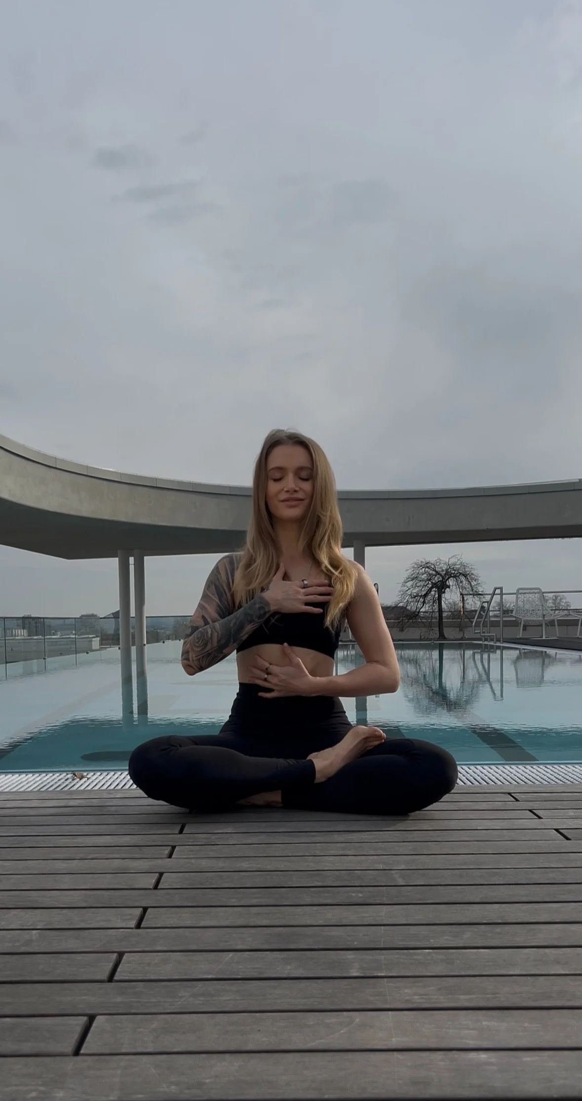 Junge Frau in Yoga-Pose am Pool auf einem Dach mit wolkigem Himmel, Himmel, Pool, Holzdeck, Outdoor-Terrasse, Bäume im Hintergrund.