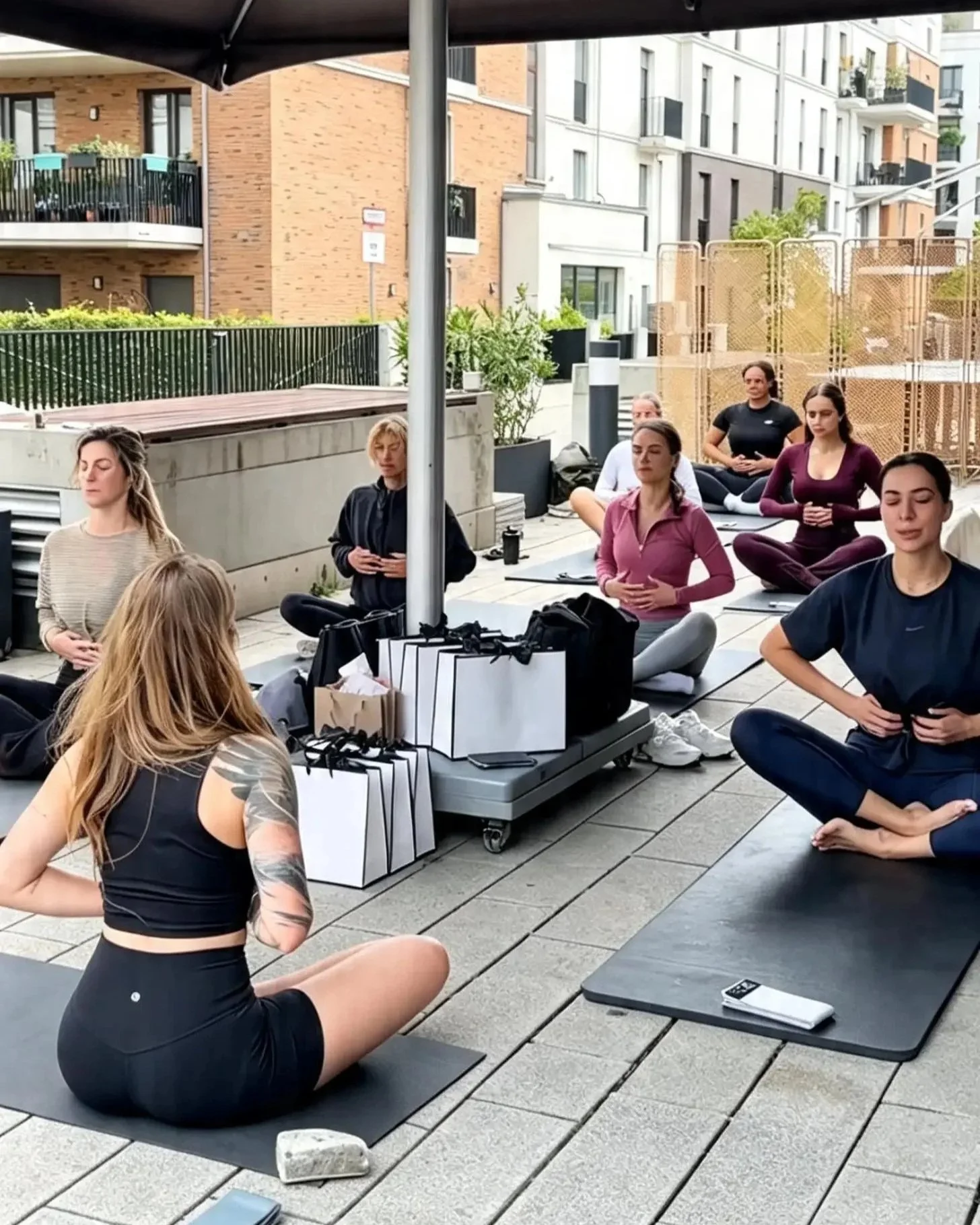 Menschen machen Open Air Pilates auf einem Balkon oder einer Terrasse, sitzen gekreuzt mit geschlossenen Augen, während eine Frau vorne zu ihnen spricht oder anleitet.