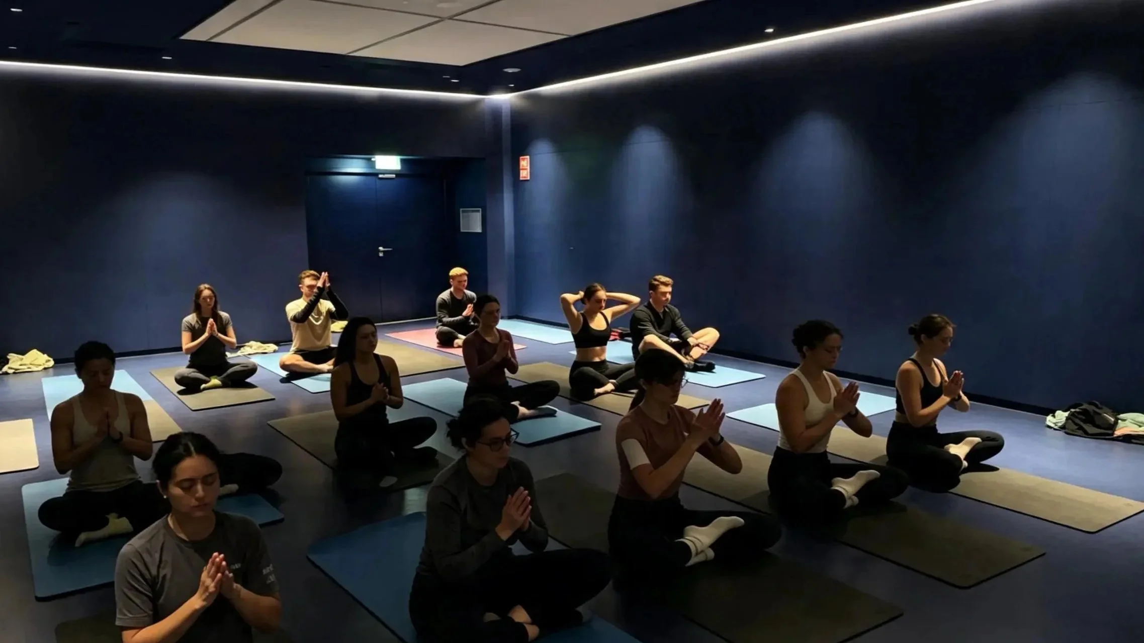 Gruppe von Menschen, die in einem edlen Raum auf Yogamatten im Lotussitz meditieren, die Hände in Gebetshaltung, während sie die Augen schließen.