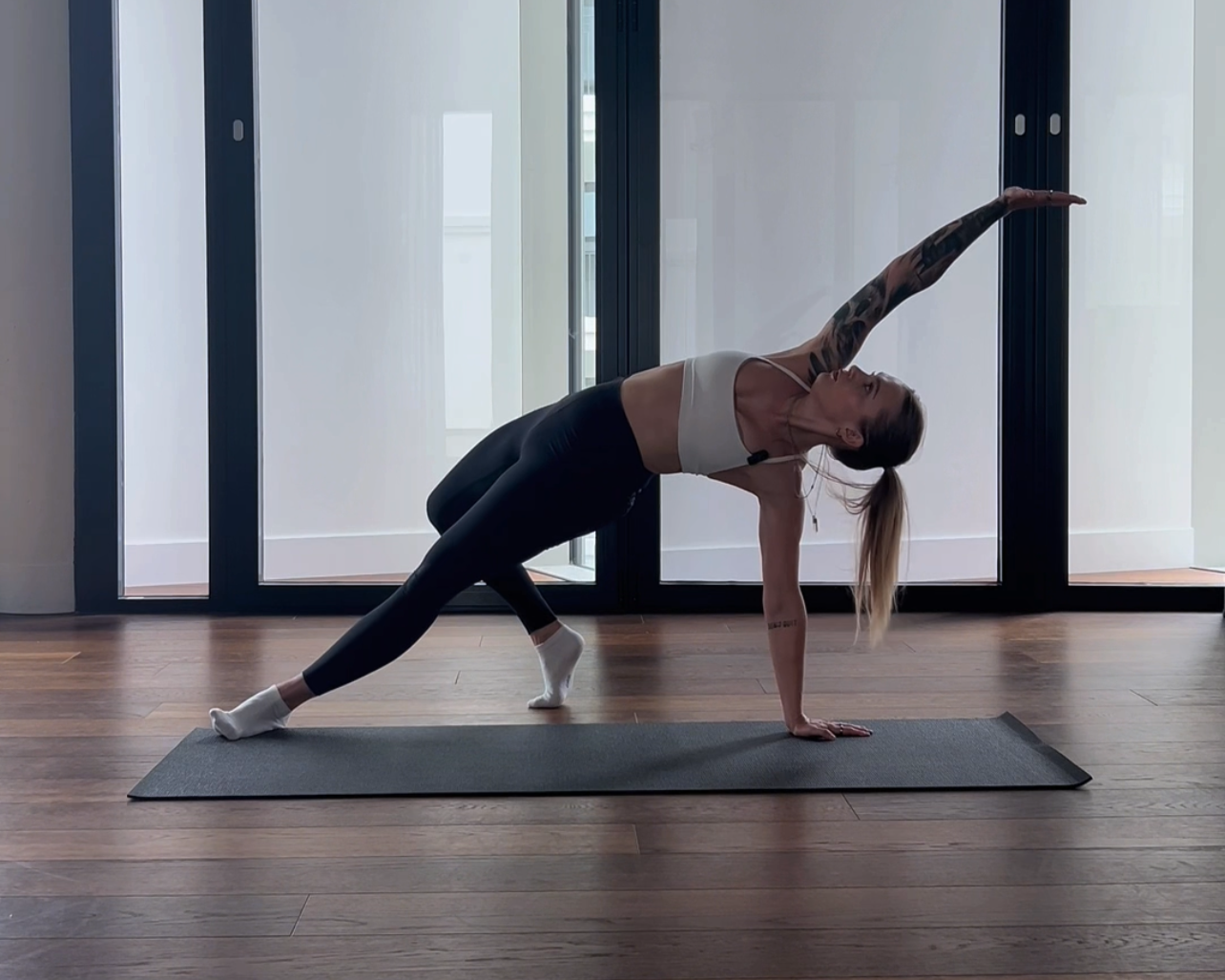Frau macht Yoga im Wohnzimmer, befindet sich in der Seitlage Position, streckt den Arm nach oben und hält einen Blick nach vorne.