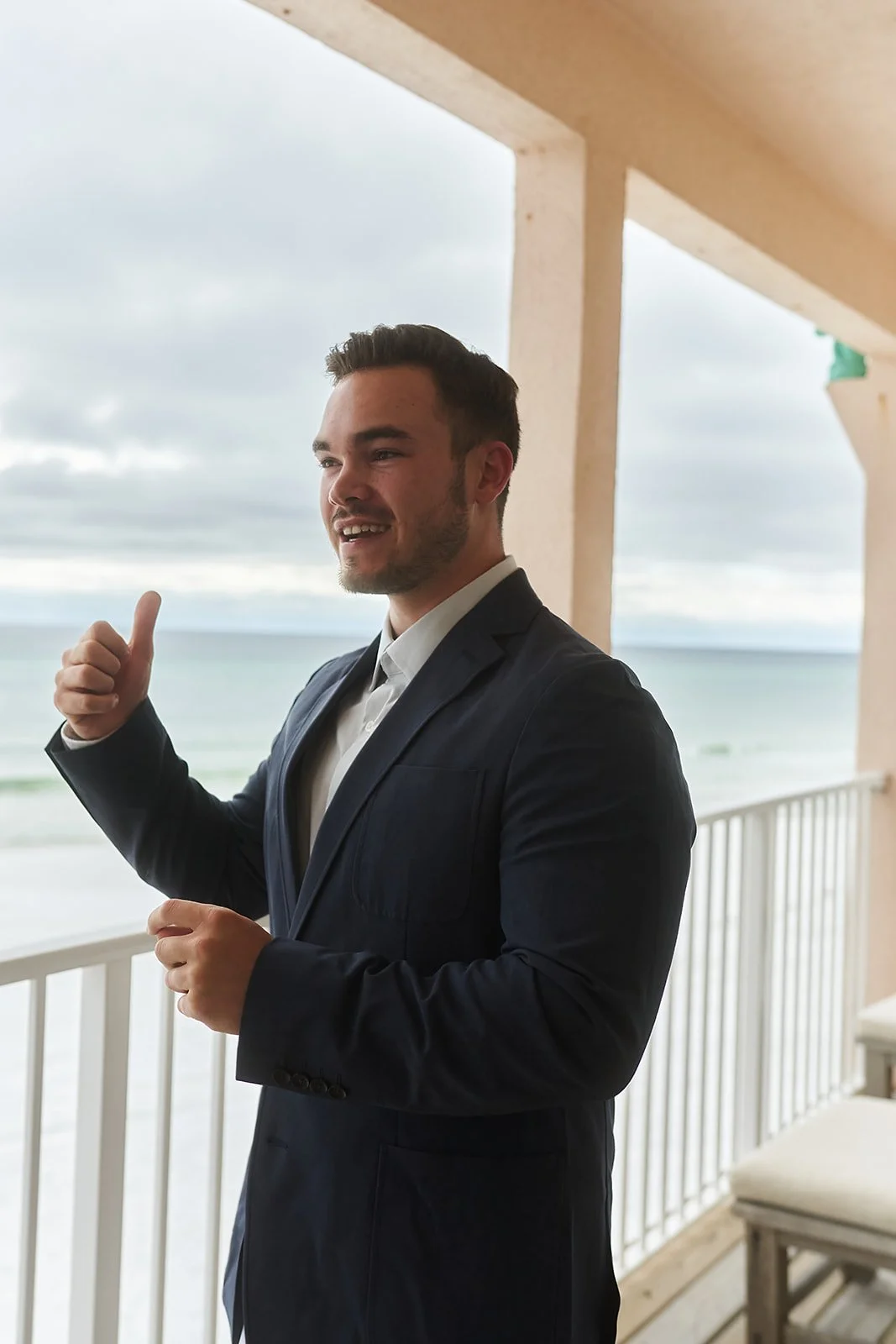 Austin Ariail in a dark suit giving a thumbs-up on a balcony overlooking the ocean, with a cloudy sky in the background.