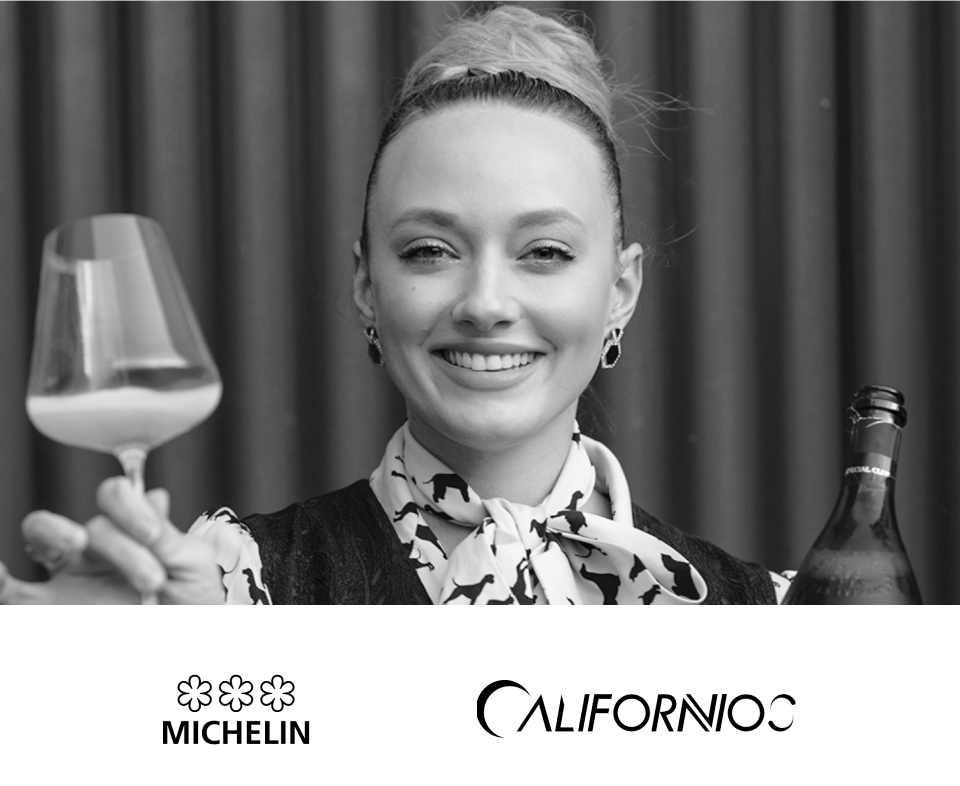 A smiling woman with her hair in a bun, wearing earrings and a patterned scarf, holding a glass of white wine in one hand and a bottle of wine in the other, standing in front of a curtain.