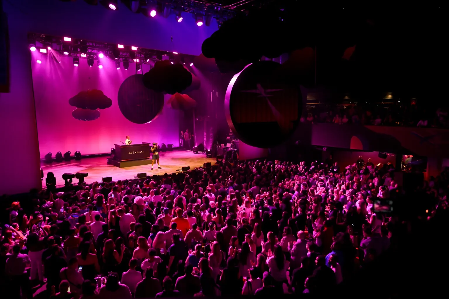Crowd watching a live performance on stage with purple lighting, a DJ booth, and decorative hanging clouds in a large indoor venue.