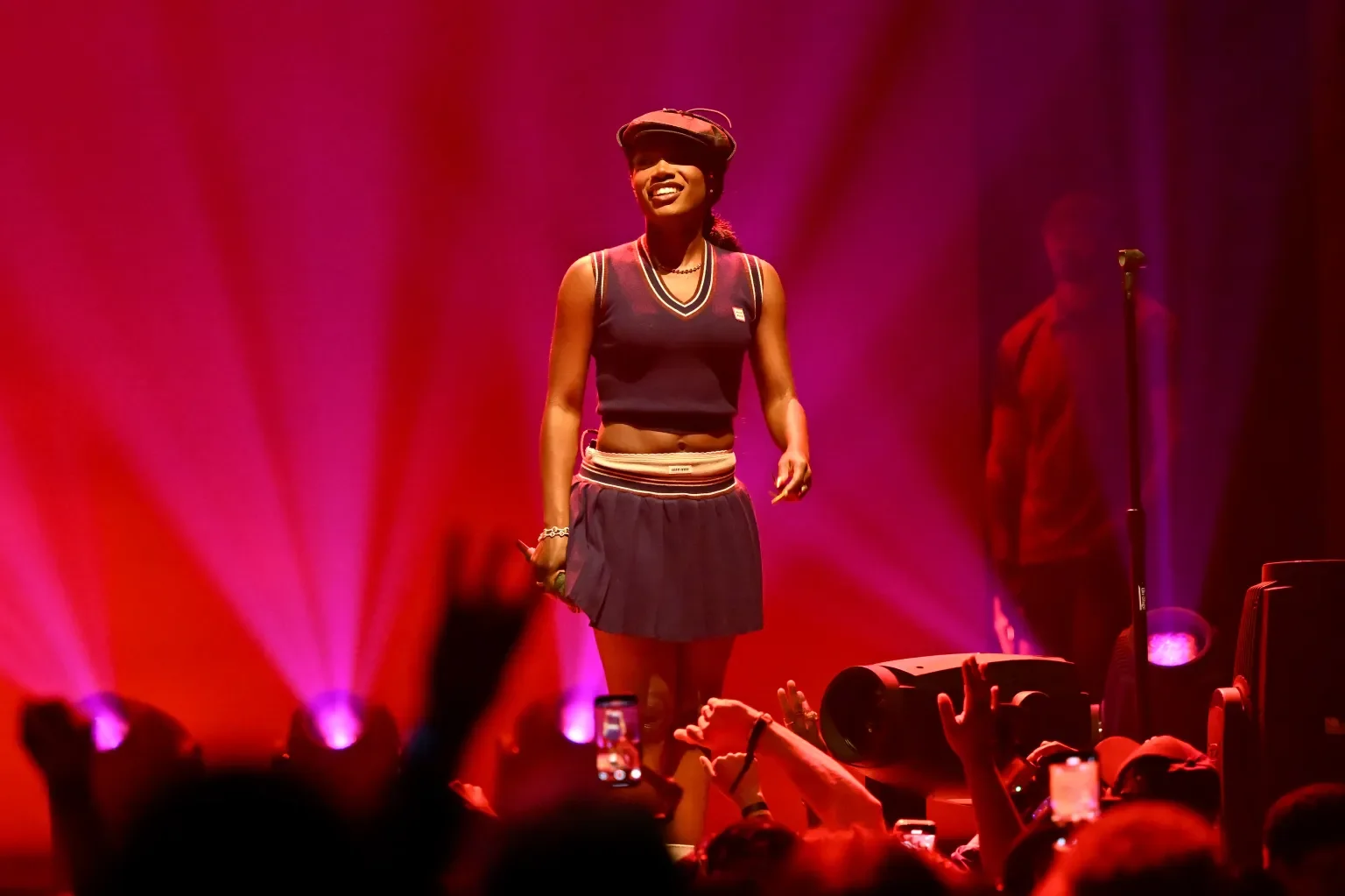 Performer on stage in colorful lighting, wearing a sporty outfit and smiling at audience, with fans taking photos.