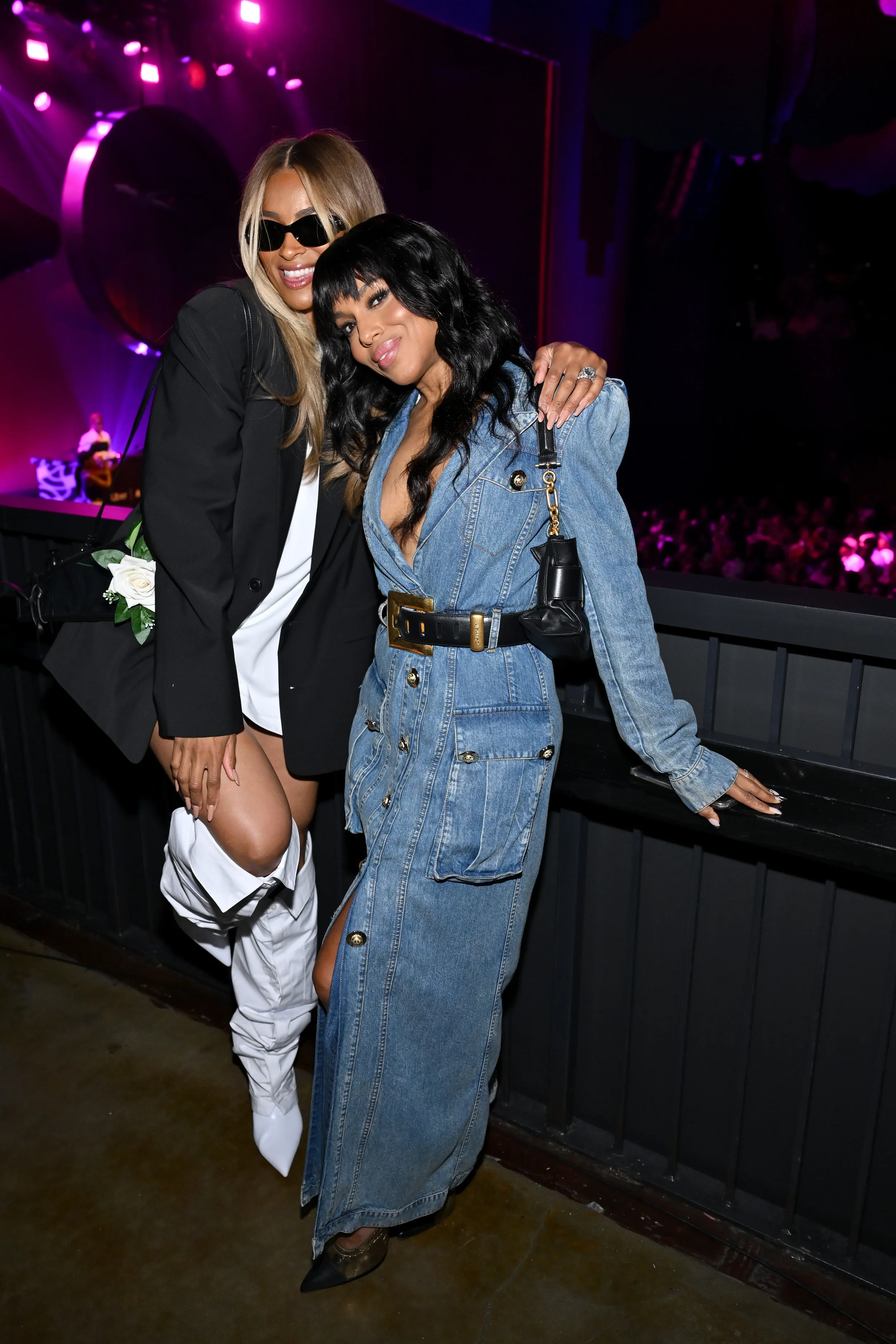 Two women pose together at a concert or event with purple stage lighting in the background. One woman has blonde hair, wears sunglasses, a black blazer, and white pants. The other woman has black hair, wears a denim dress, and carries a small black purse. They are smiling and standing close to each other.